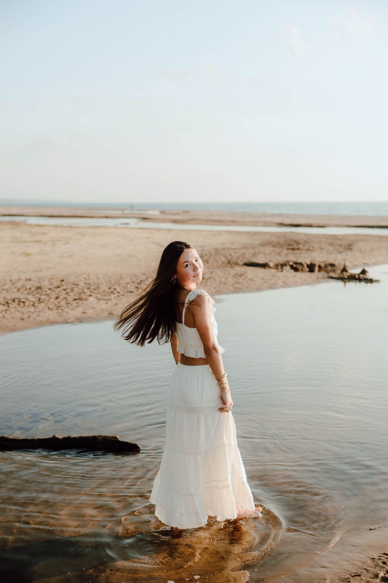 Woman in white dress standing in shallow beach water, hair flowing, with sandy shore and ocean in the background.