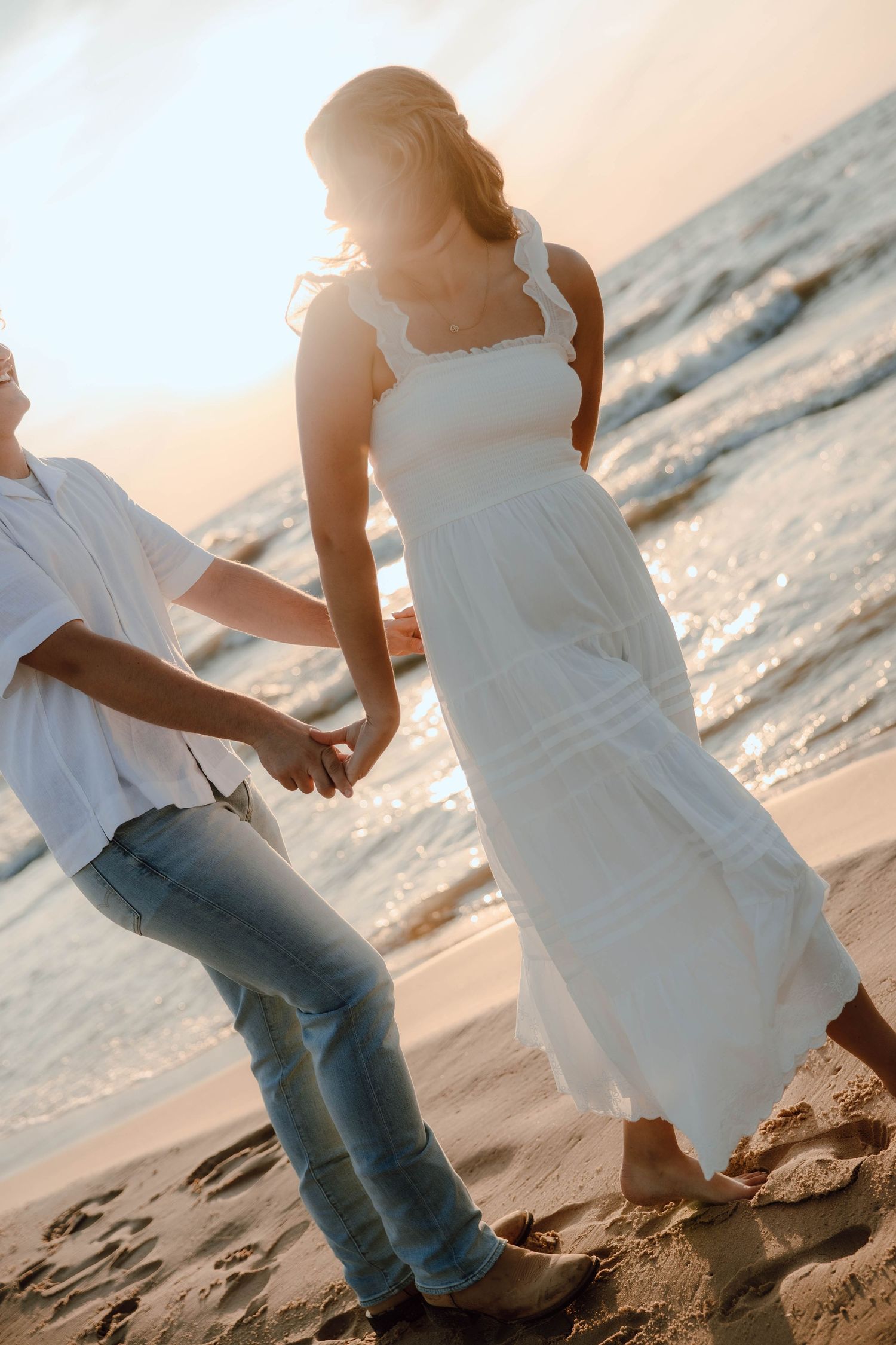 Couple holding hands walking on a sandy beach at sunset, woman in white dress, man in jeans and white shirt.