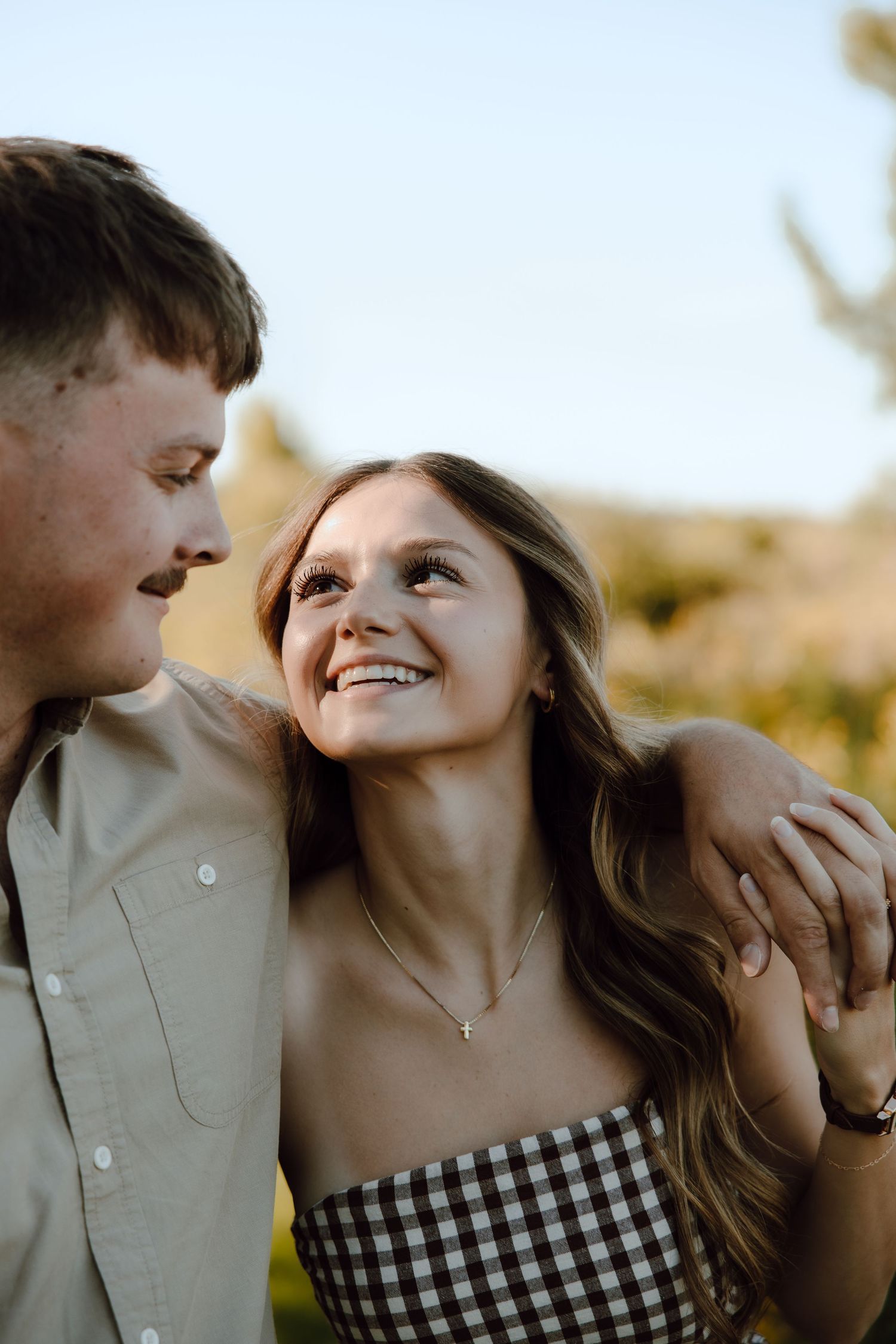 A young couple smiles warmly at each other outdoors, the woman in a plaid top wearing a cross necklace.