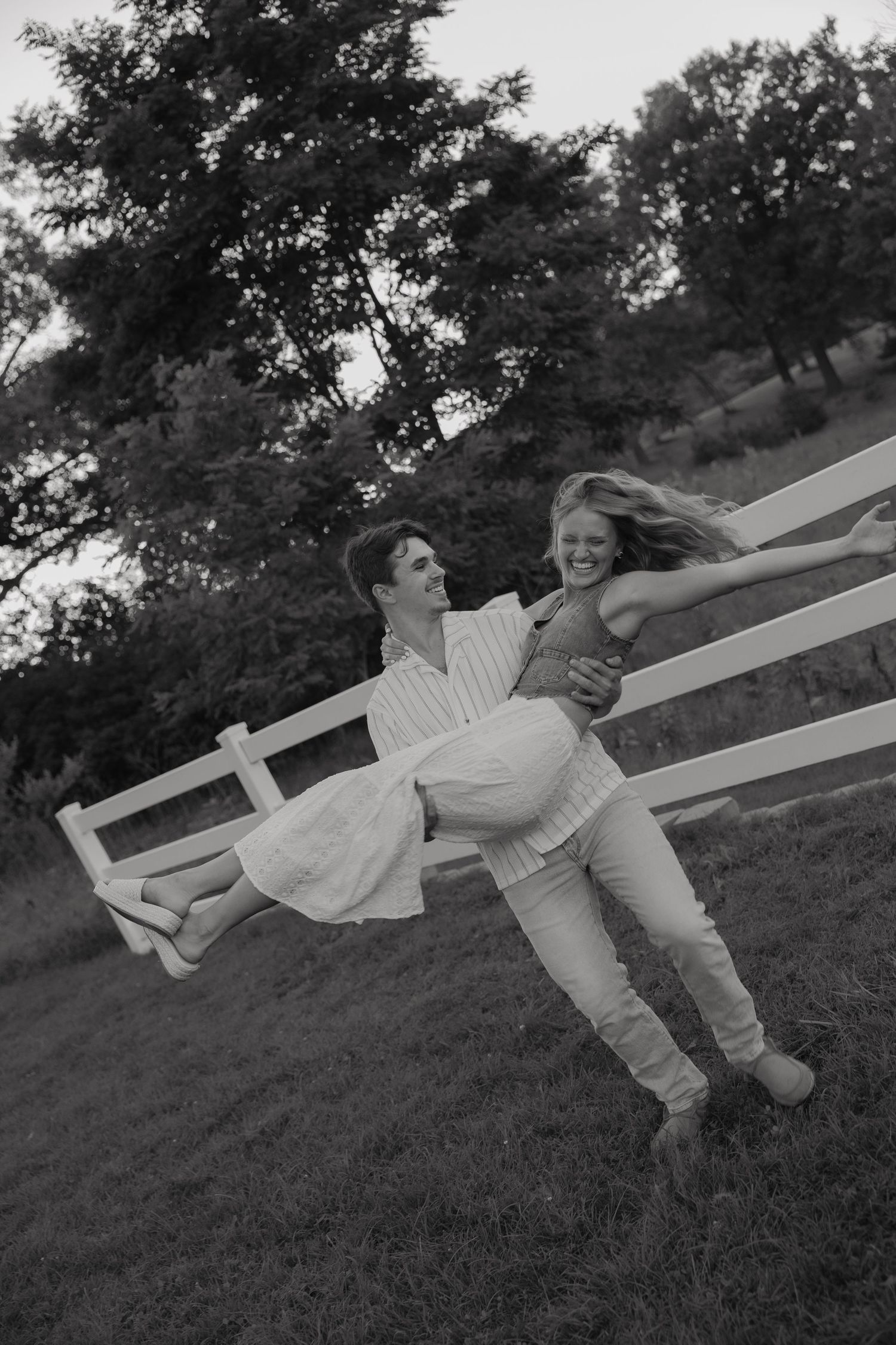 Black and white photo of a man carrying a laughing woman in his arms near a white fence outdoors.