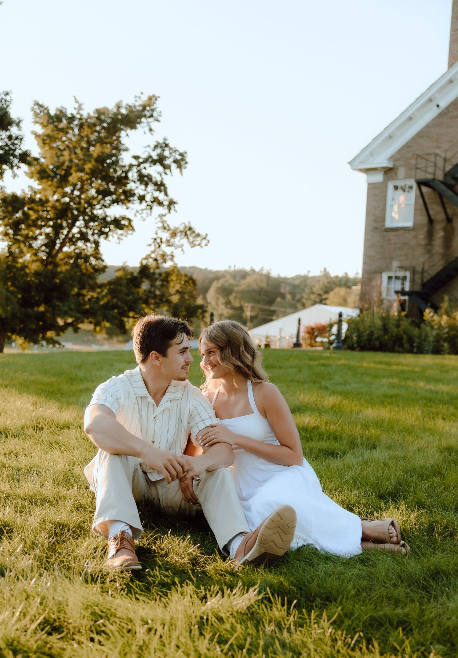 A couple sits together on green grass, smiling at each other near a brick building on a sunny day.