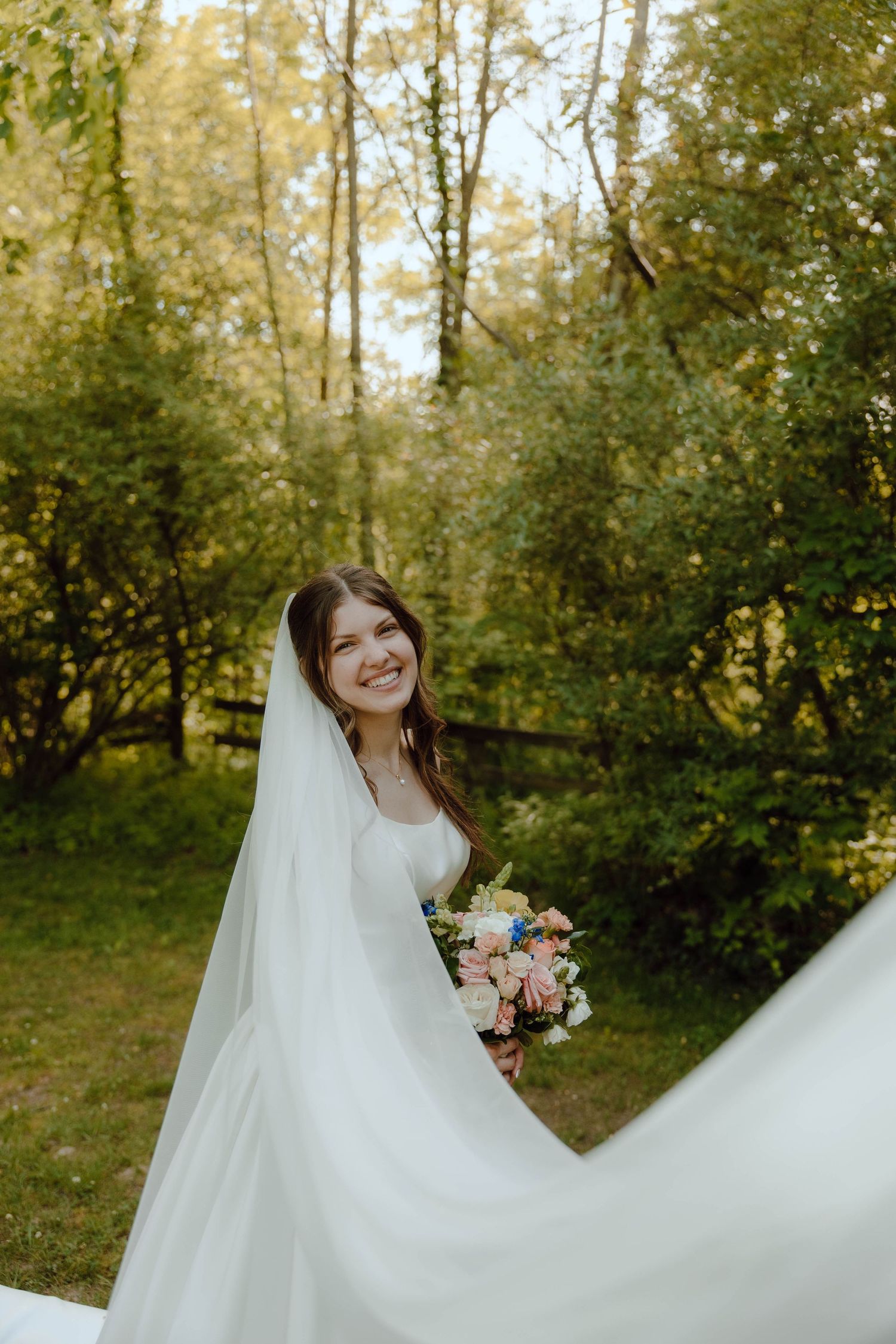 Joyful bride in white dress and flowing veil smiles while holding a pink and blue floral bouquet outdoors.