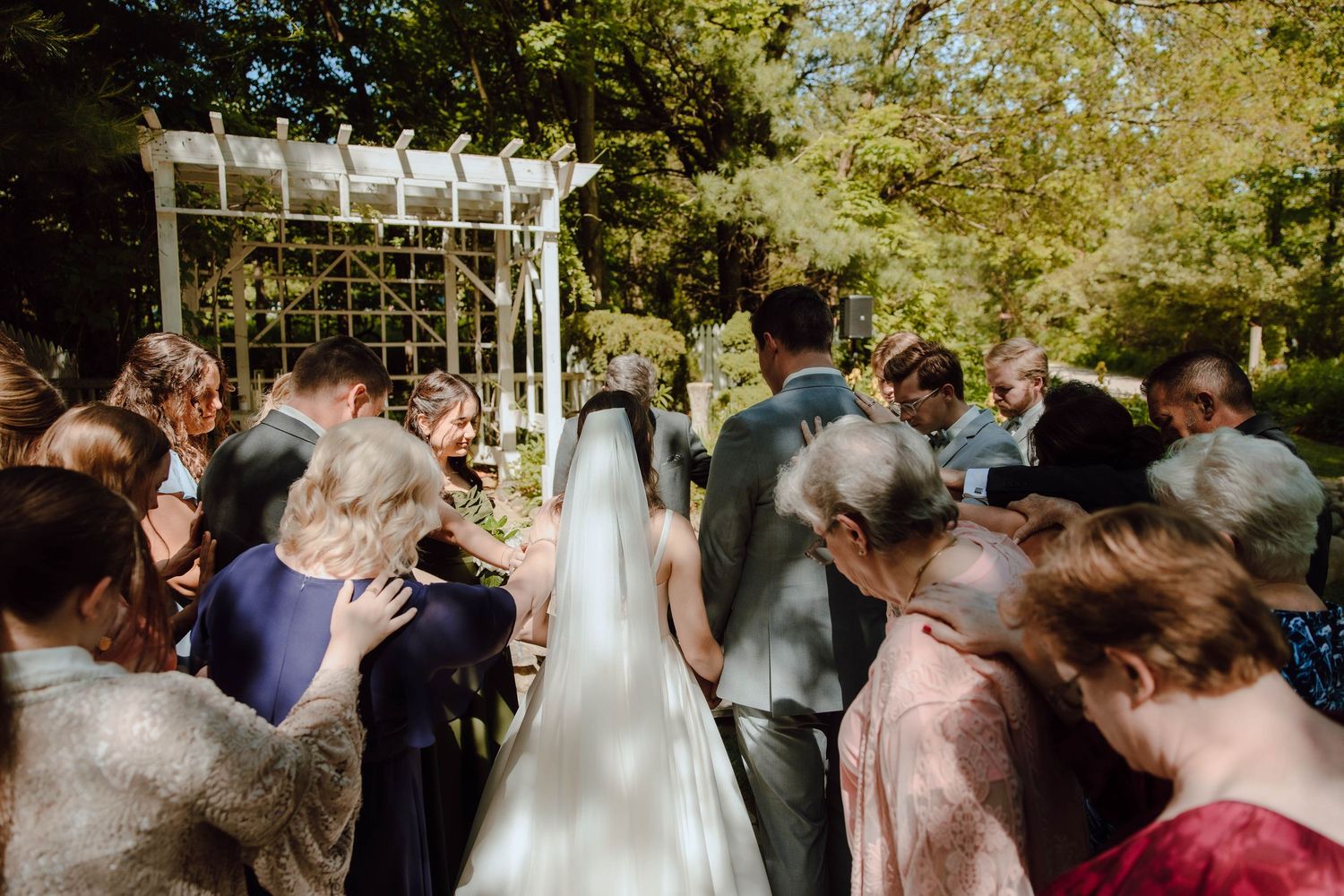 Outdoor wedding ceremony with bride in white gown and veil, guests gathered under white pergola surrounded by lush green trees.