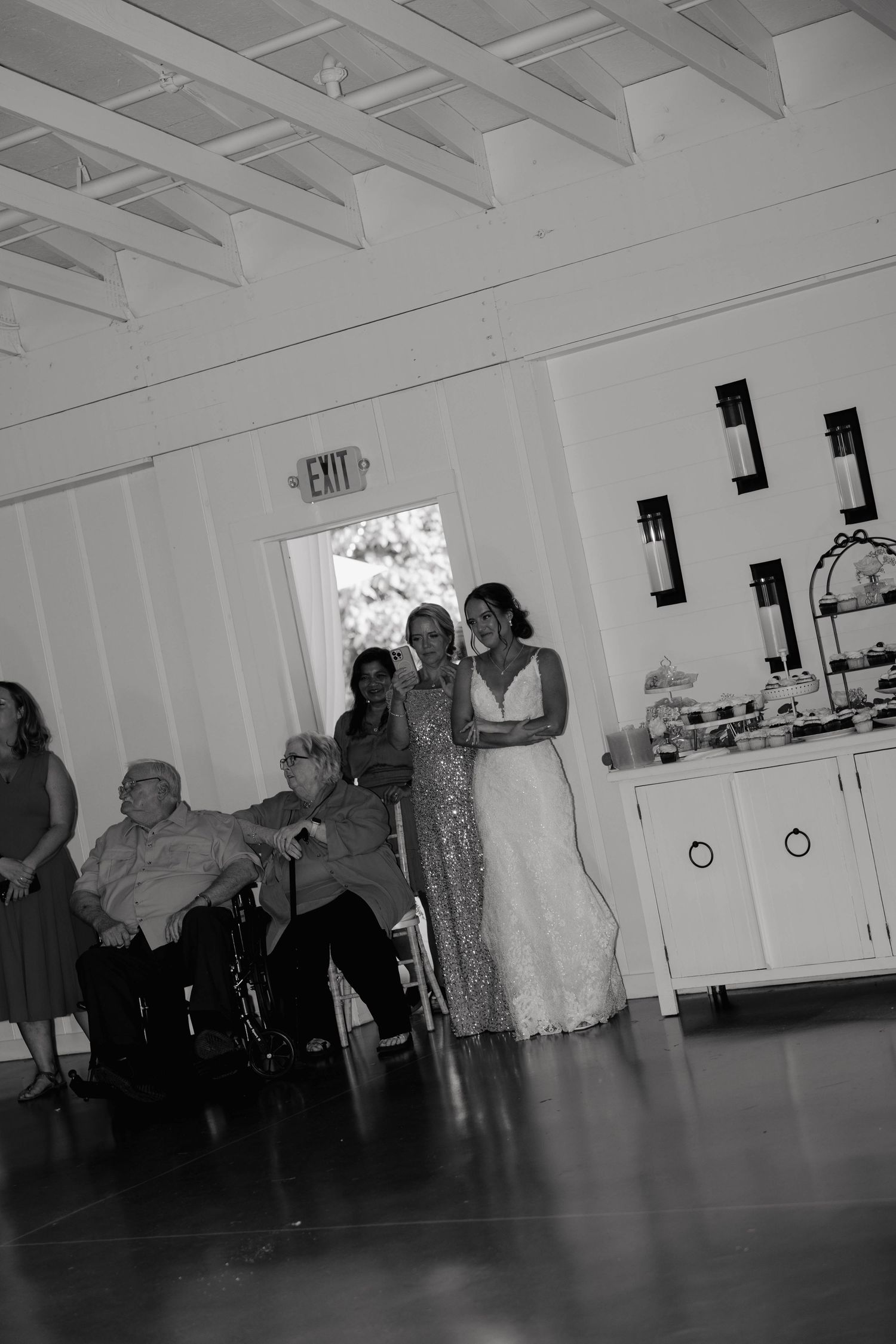 Black and white wedding reception photo showing bride in lace gown posing with guests in elegant white venue.