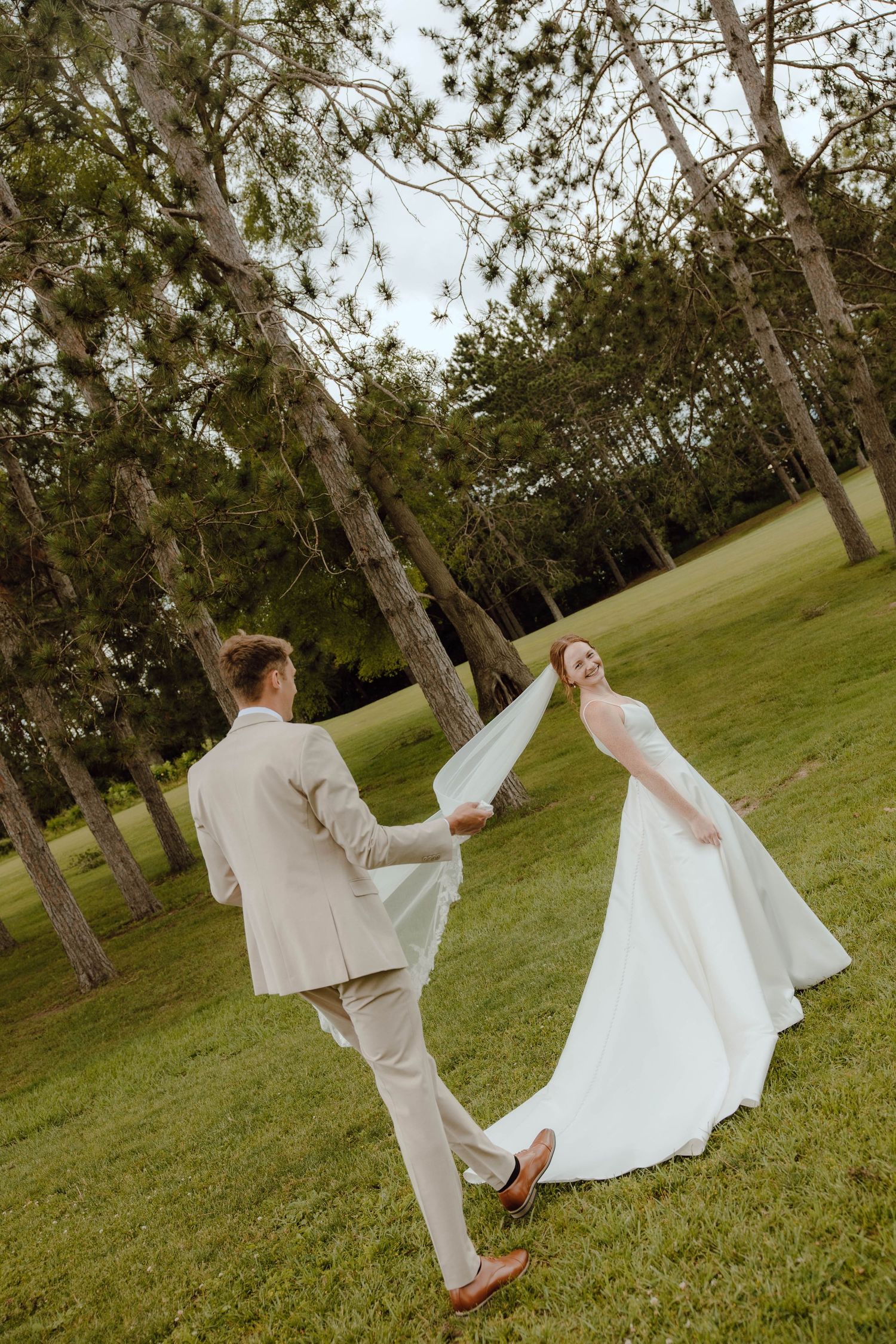 Bride and groom dancing joyfully in a lush green park, bride in white gown, groom in beige suit.