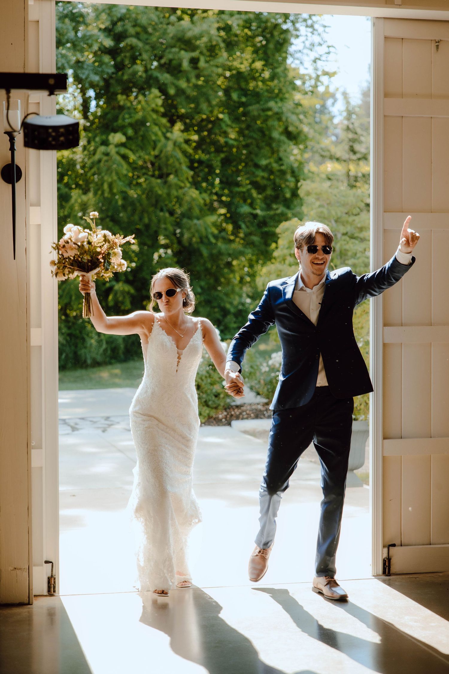 Joyful newlyweds wearing sunglasses make grand entrance through barn doors holding bouquet at wedding reception.