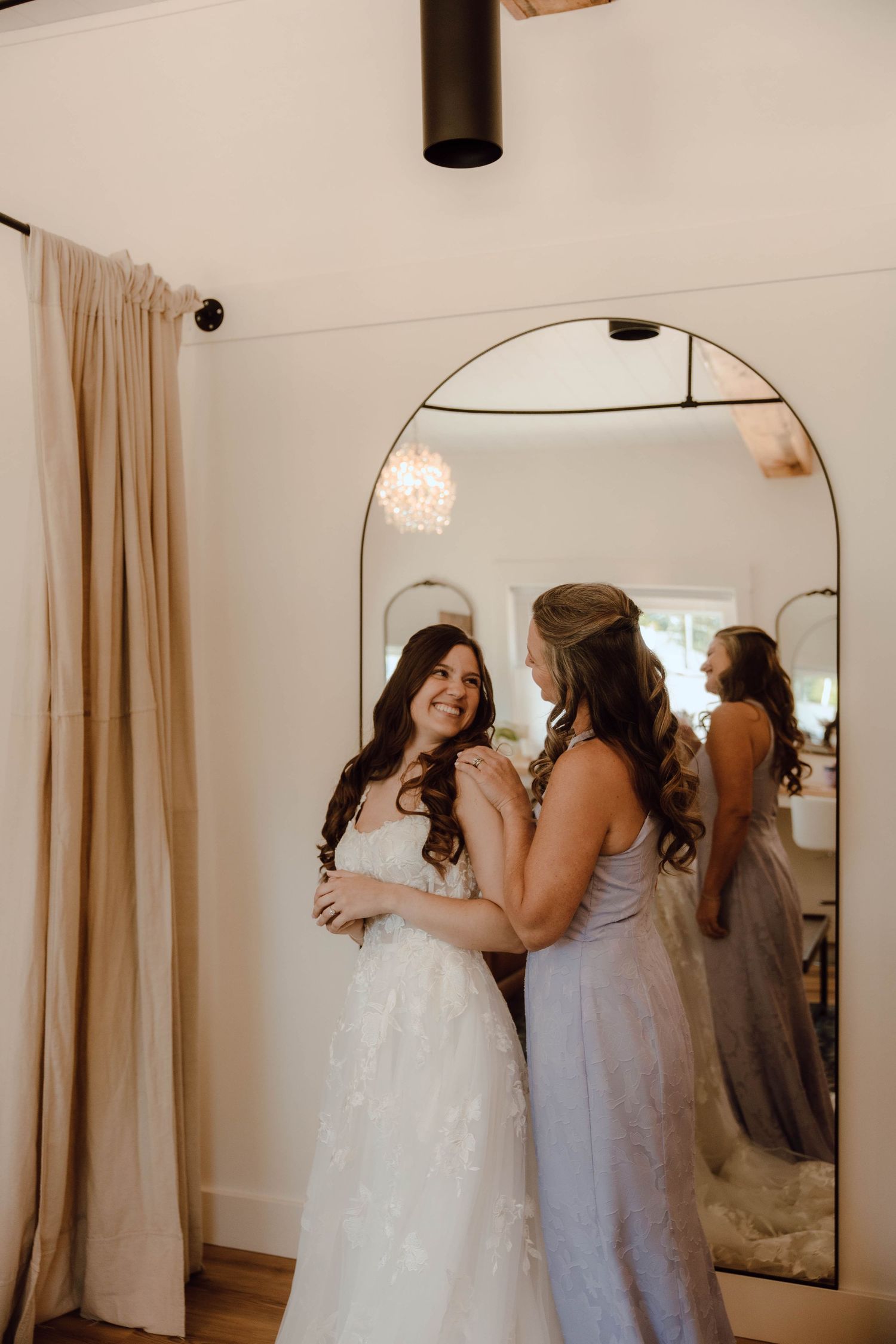 Bride in white lace gown smiling with bridesmaid in lavender dress, standing before arched mirror in bridal suite.