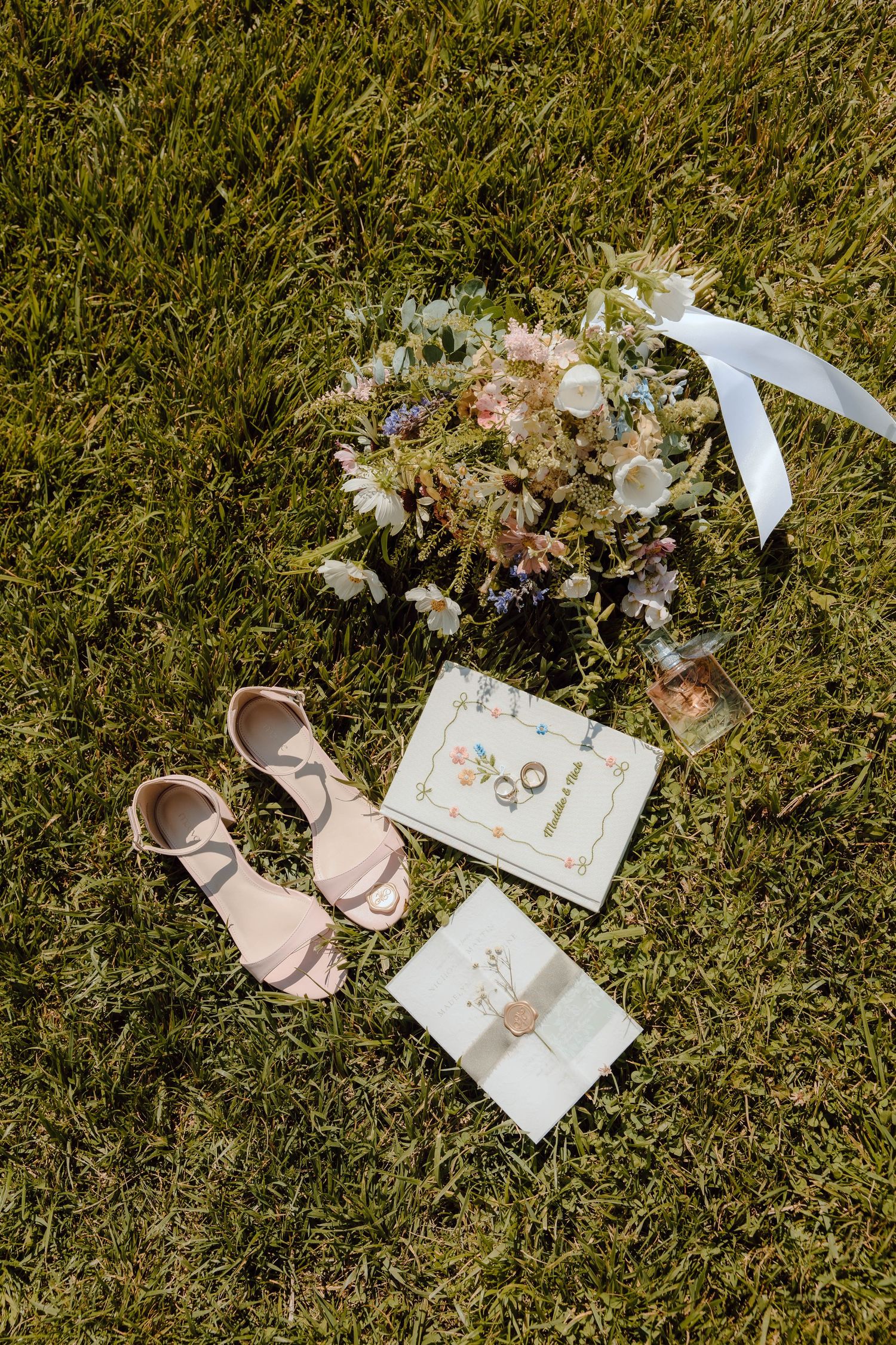 Flat lay of blush heels, floral bouquet, wedding stationery, and perfume on green grass.