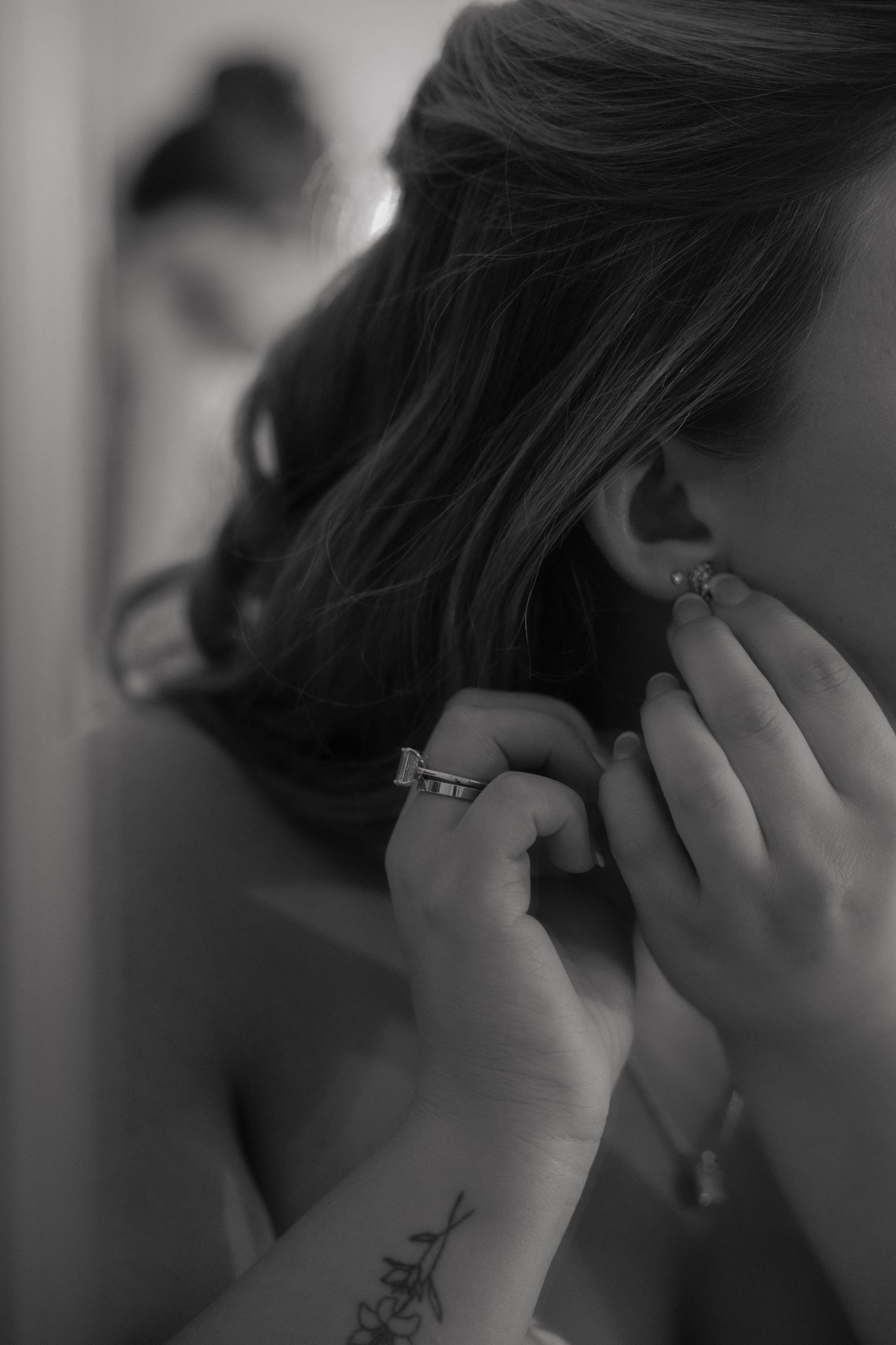 Black and white photo of a bride putting on a diamond earring with a tattoo visible on her wrist.