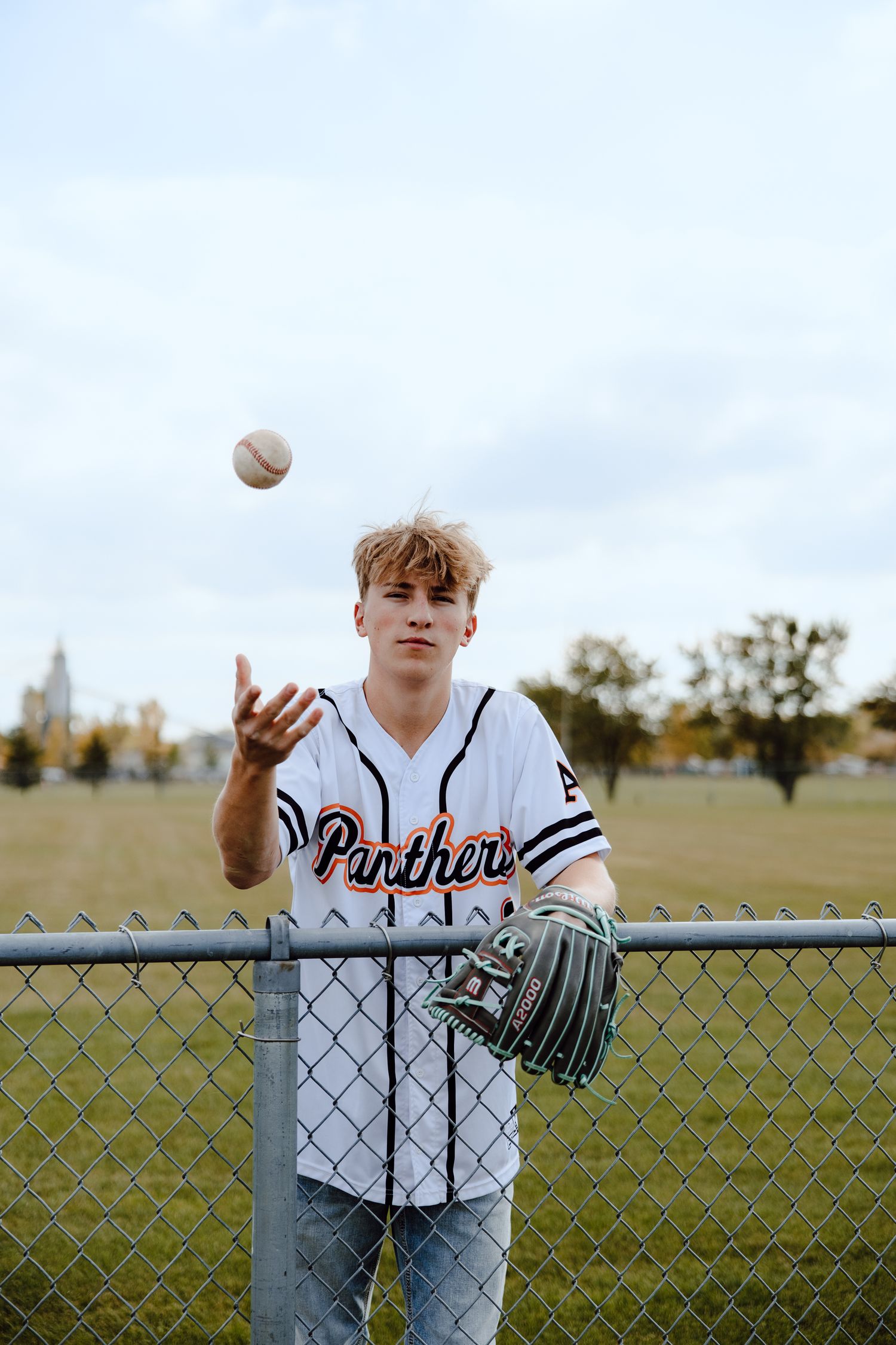 Baseball player in Panthers uniform tossing ball while leaning on chain link fence.