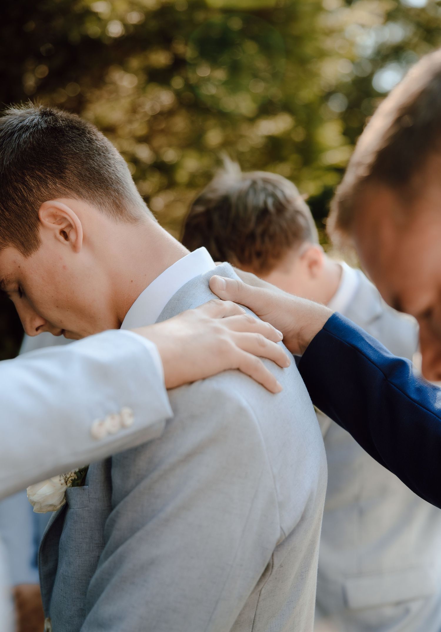 Groomsmen in light blue suits placing hands on each other's shoulders during an outdoor wedding ceremony.