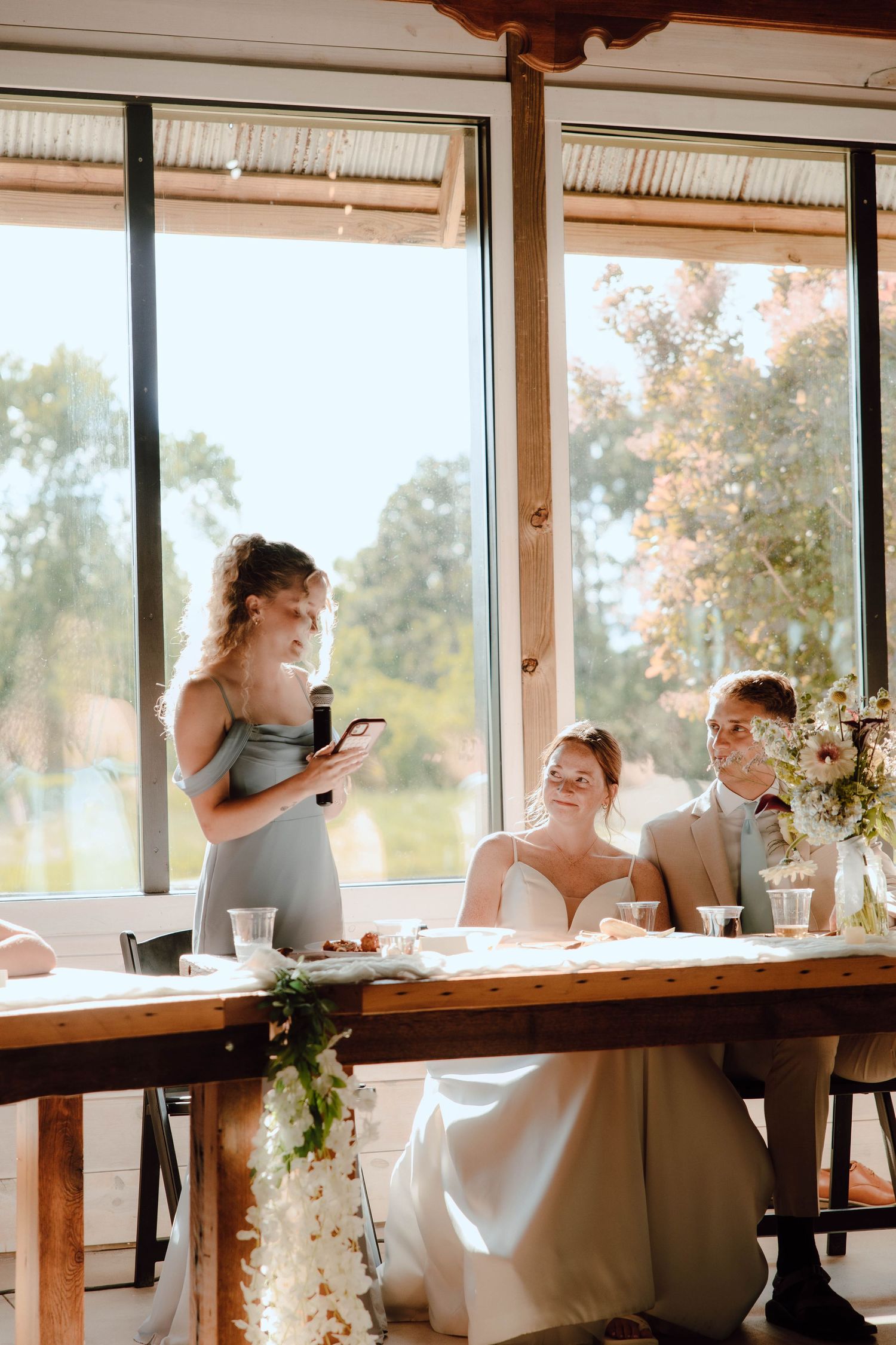Bridesmaid giving a heartfelt speech at a rustic wedding reception while the bride and groom sit at the head table.