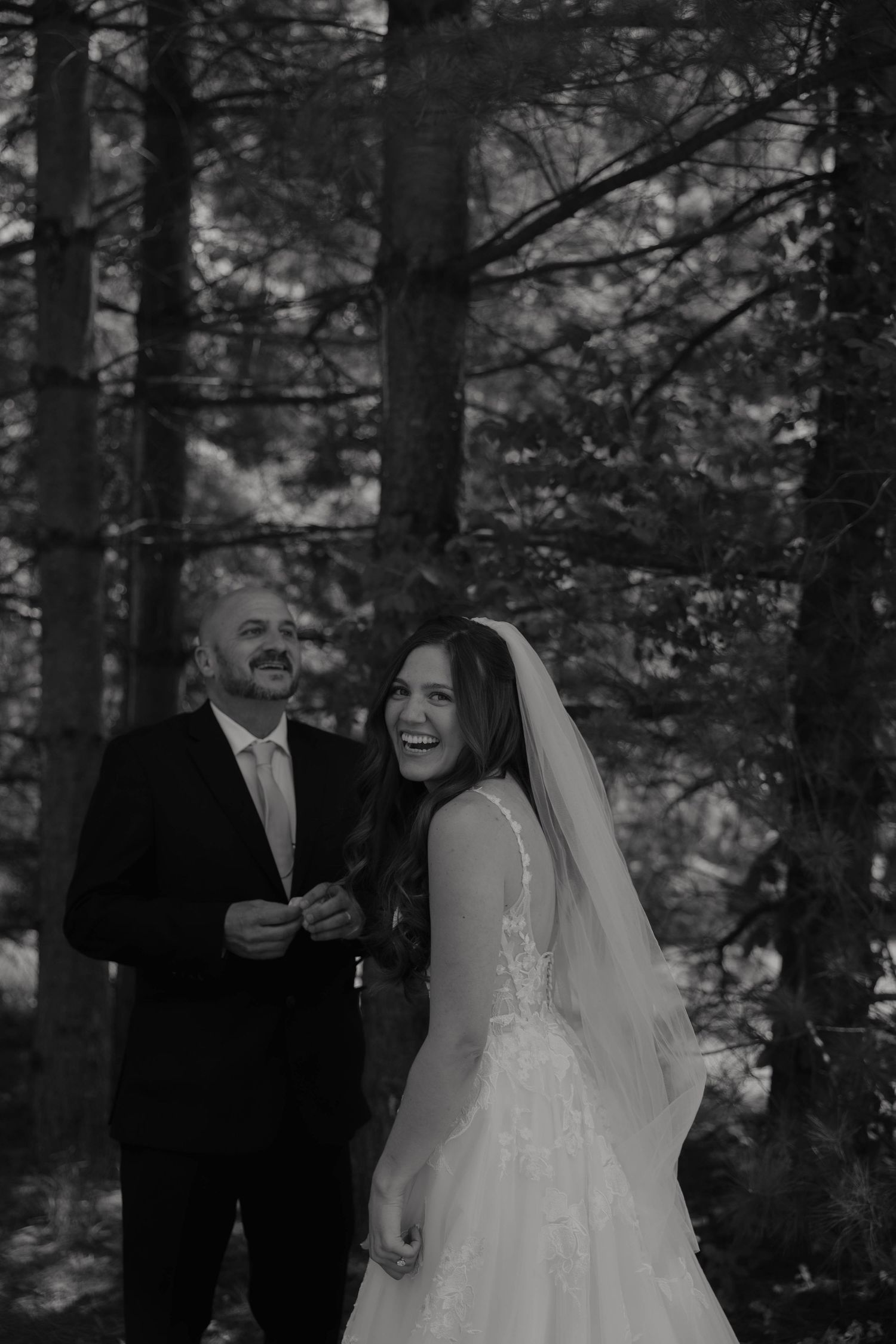 Bride in lace veil laughs joyfully with groom among tall trees in black and white forest wedding photo.