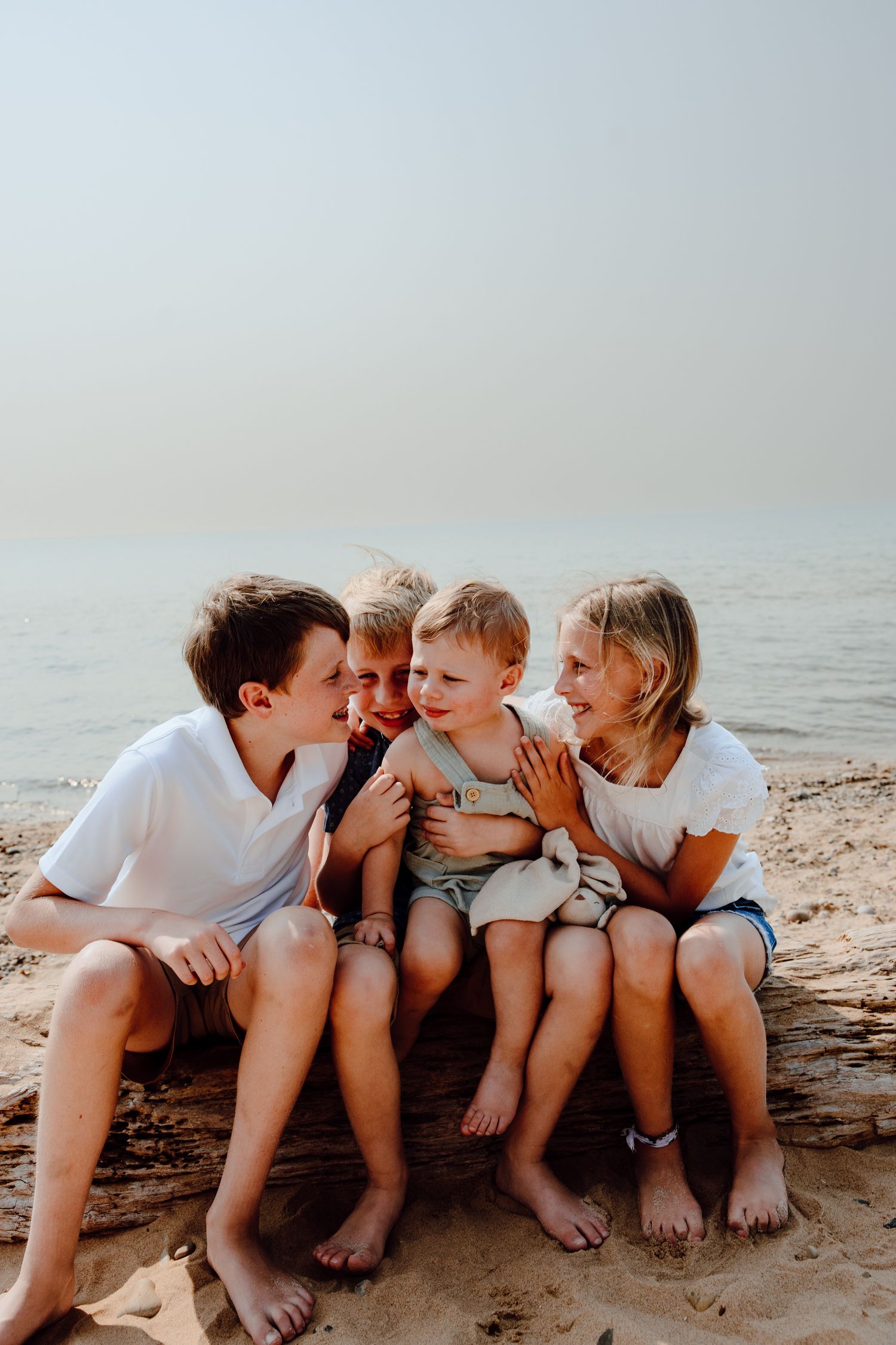 Children sit in a row on a beach log sharing secrets and laughing together.