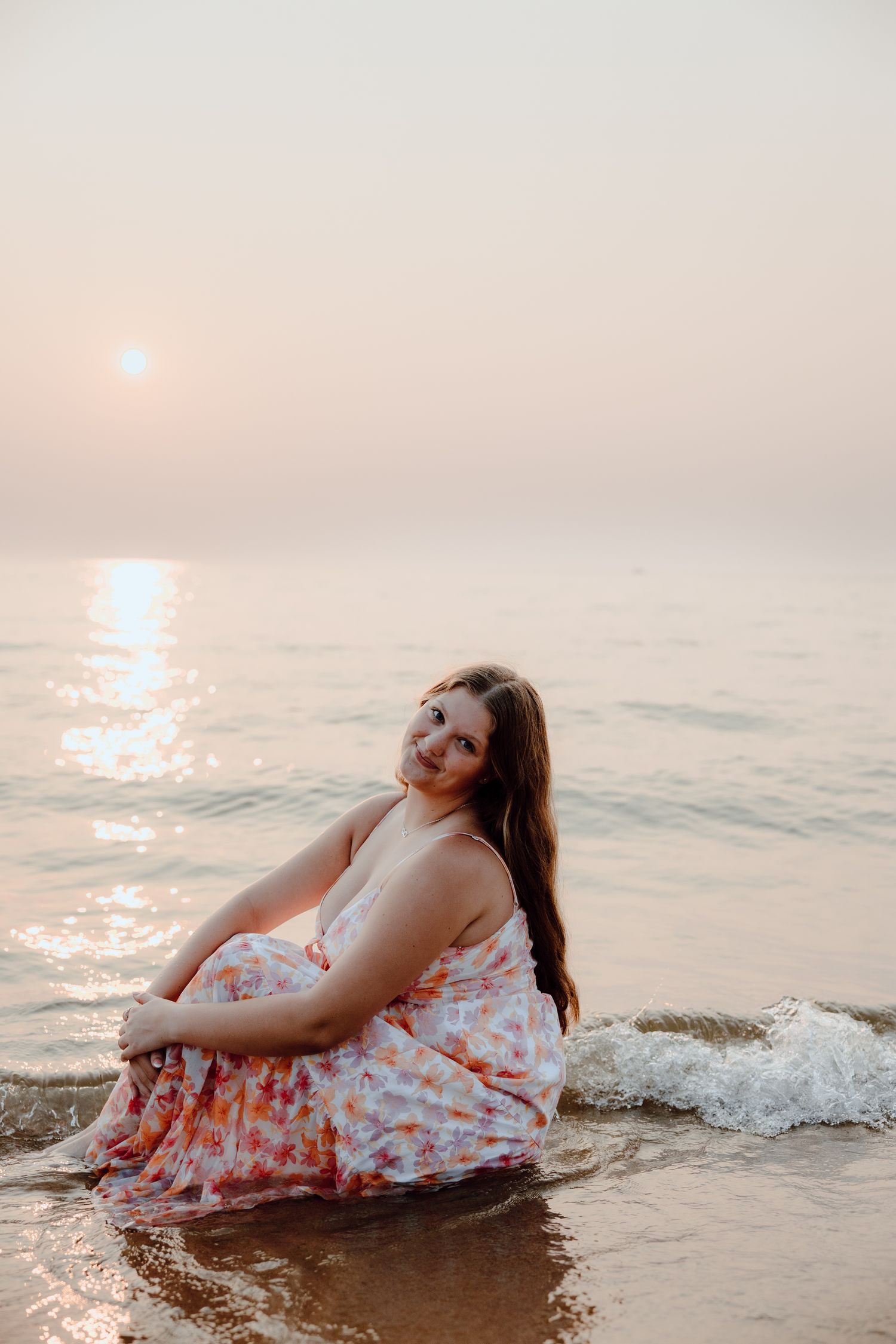 Person in floral dress sits in shallow ocean waves during peaceful sunset.