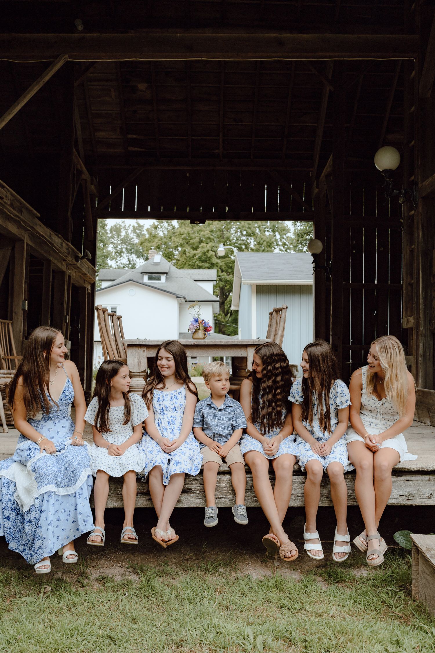 A group sits in a row under a rustic wooden barn overhang wearing matching blue and white dresses.