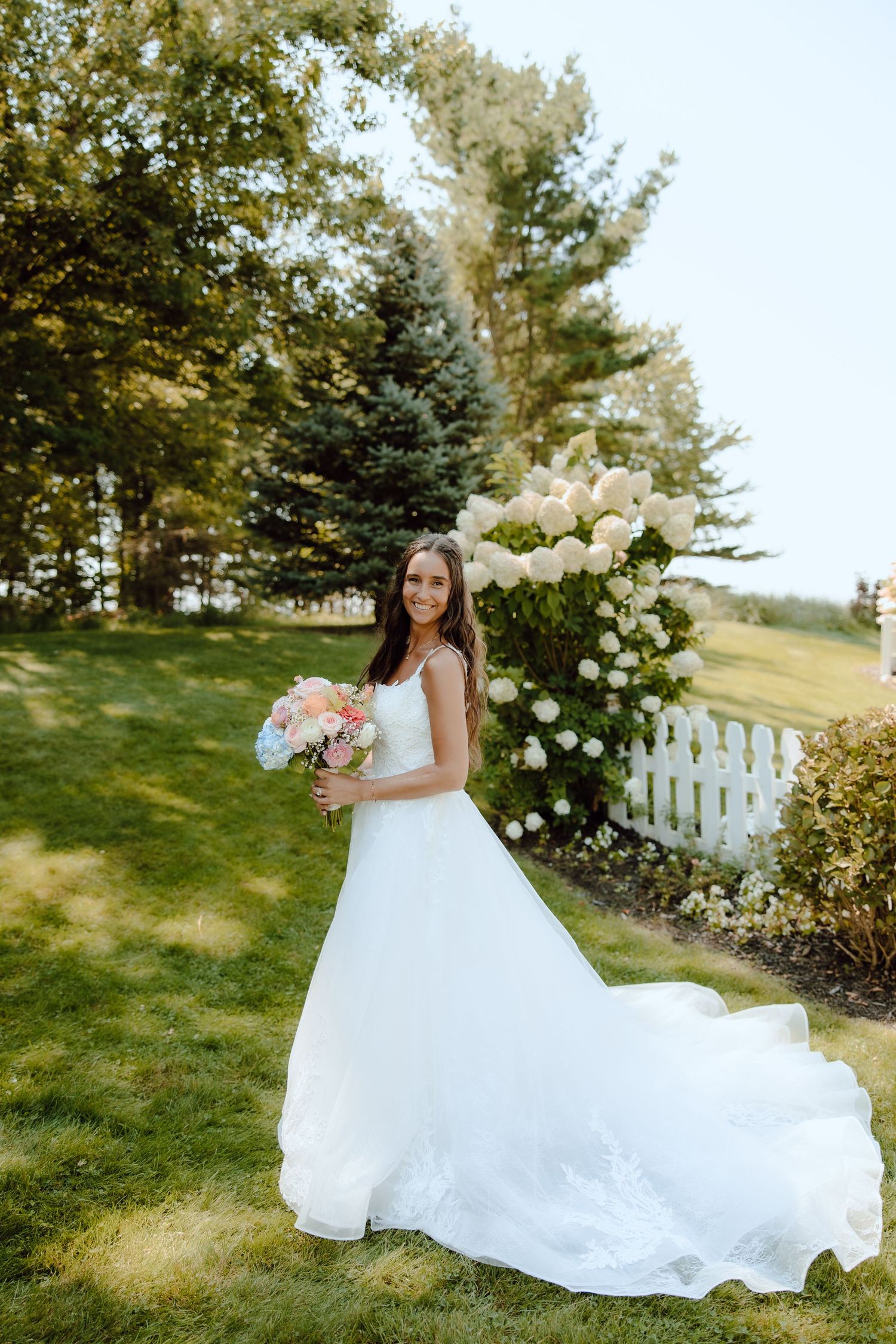 Bride in white gown holding pink bouquet poses in lush garden with white hydrangeas and picket fence.