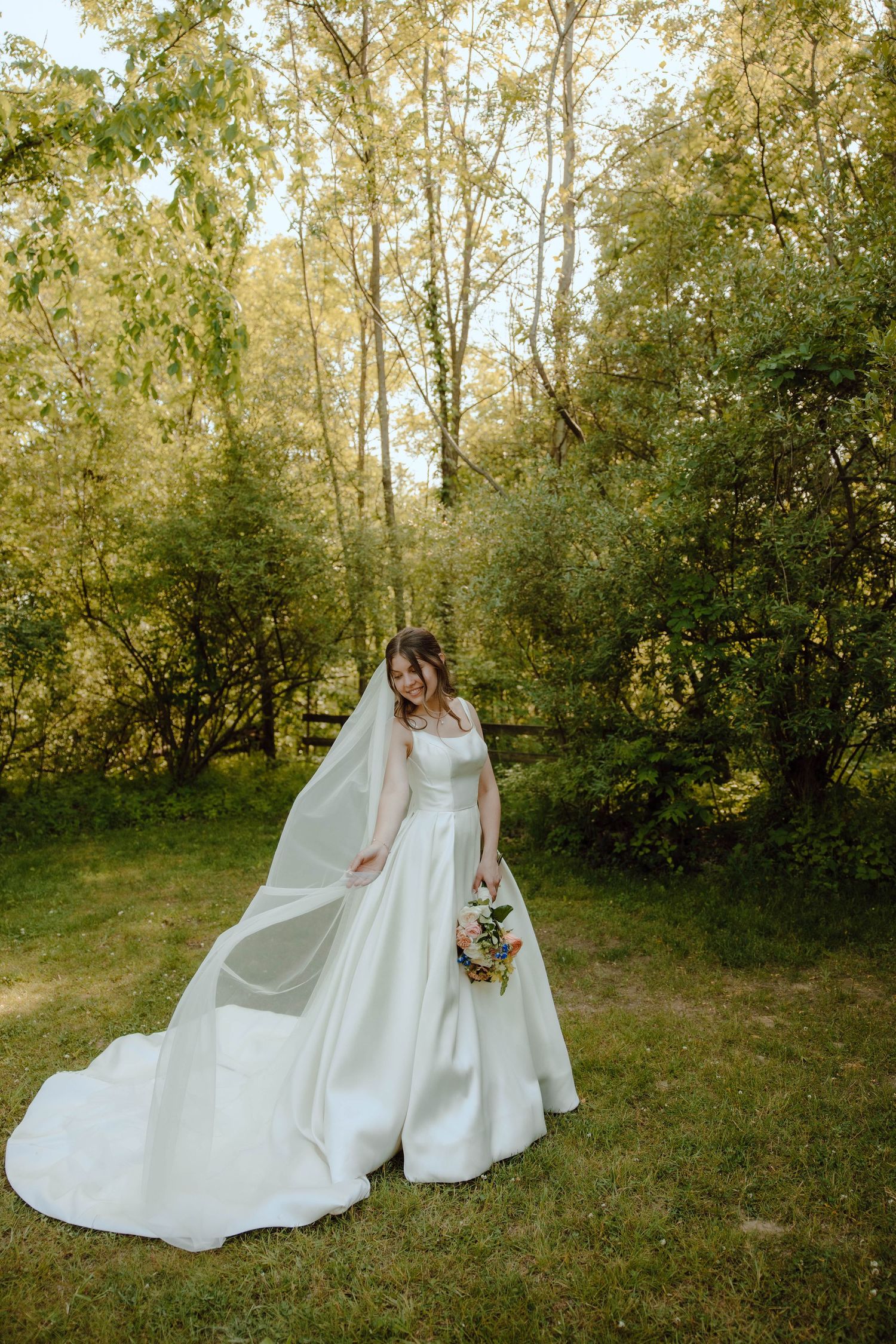Elegant bride in a white ball gown with a long flowing veil holds a bouquet in a lush green garden.