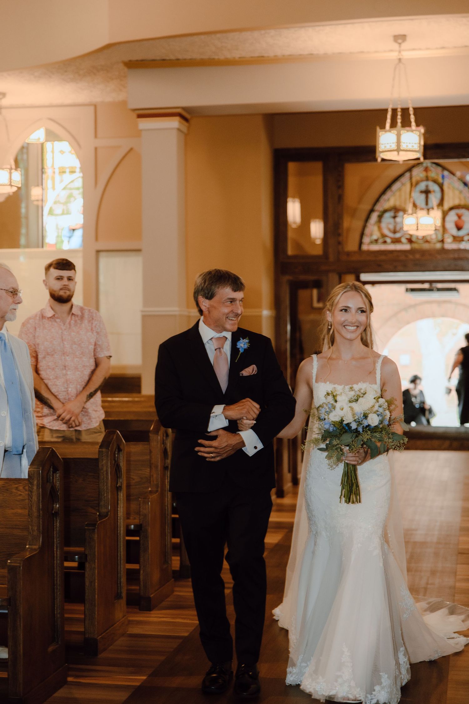 Smiling bride walks down church aisle with father holding white and blue bouquet.