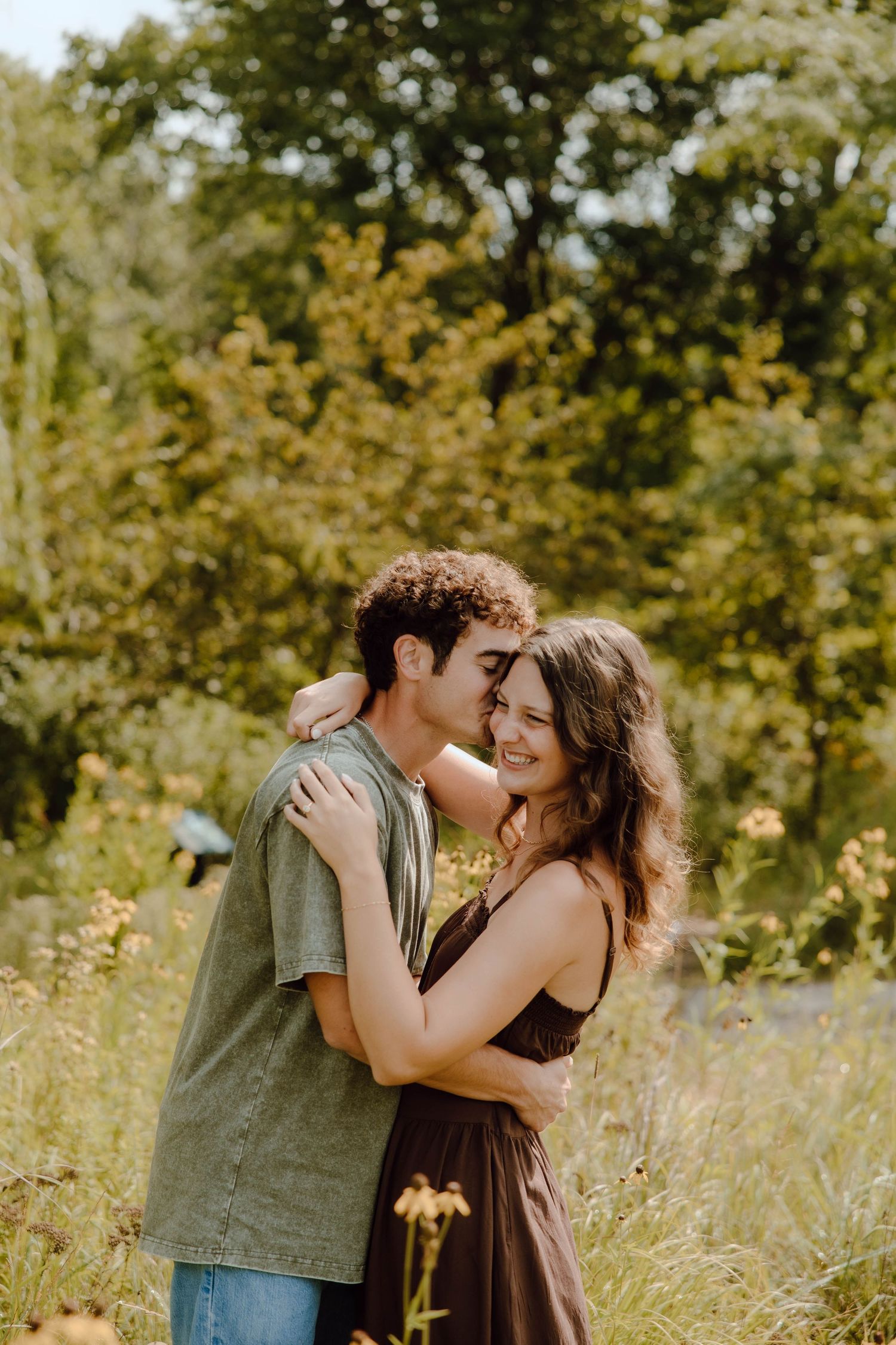 A young couple embraces and shares a tender moment in a lush green meadow with wildflowers on a sunny day.