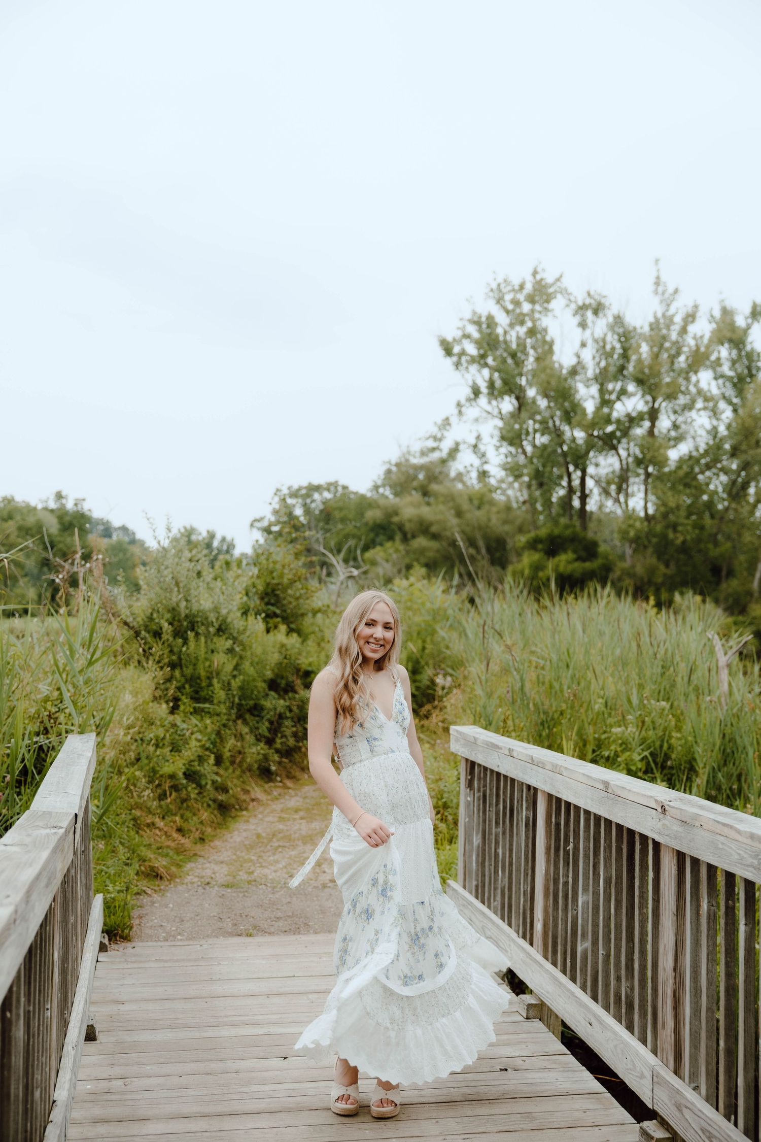 Smiling blonde woman in white floral maxi dress twirling on wooden boardwalk surrounded by lush greenery.
