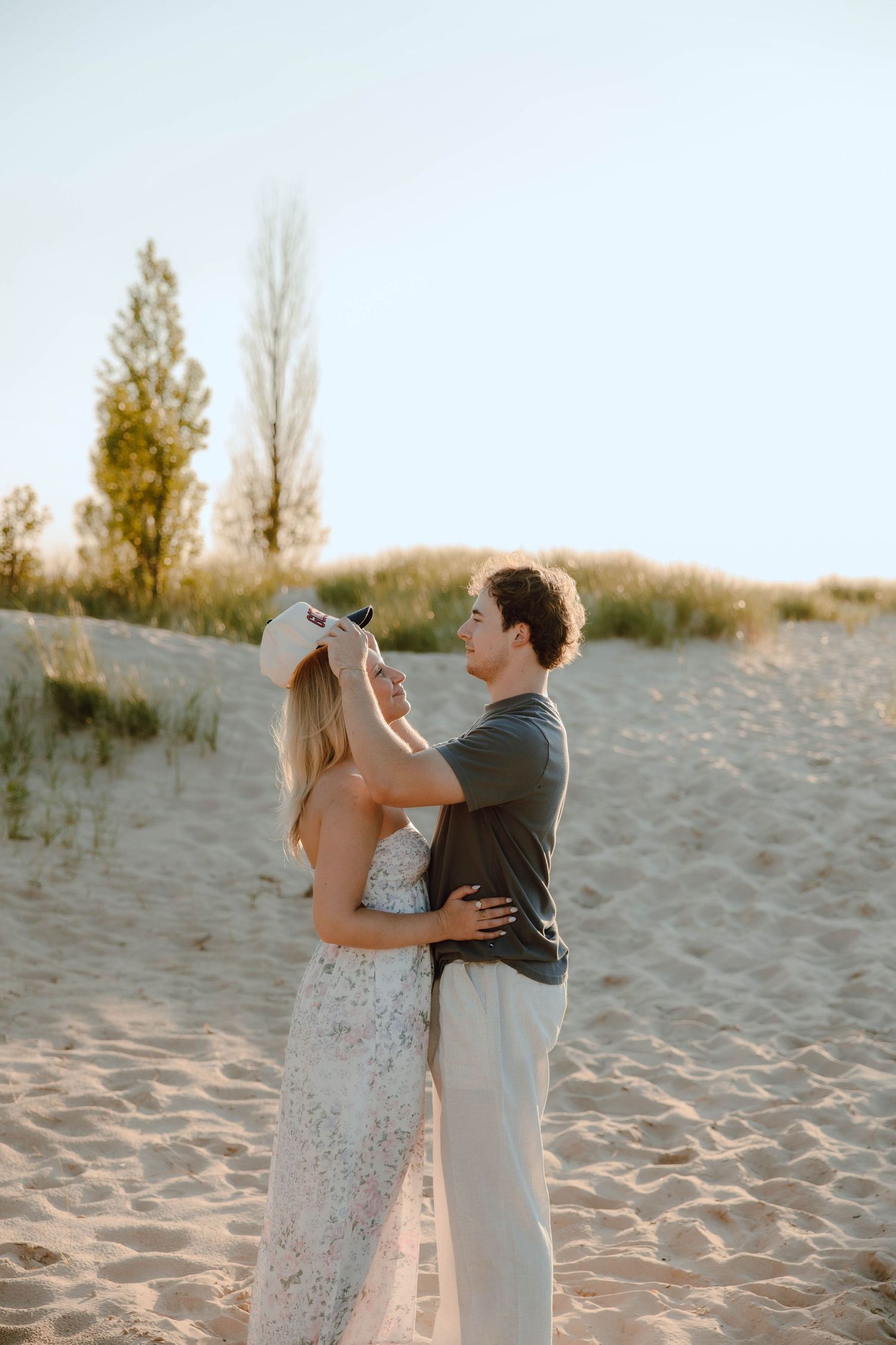 A couple shares a romantic moment on sandy dunes, as the man adjusts the woman's hat at golden hour.