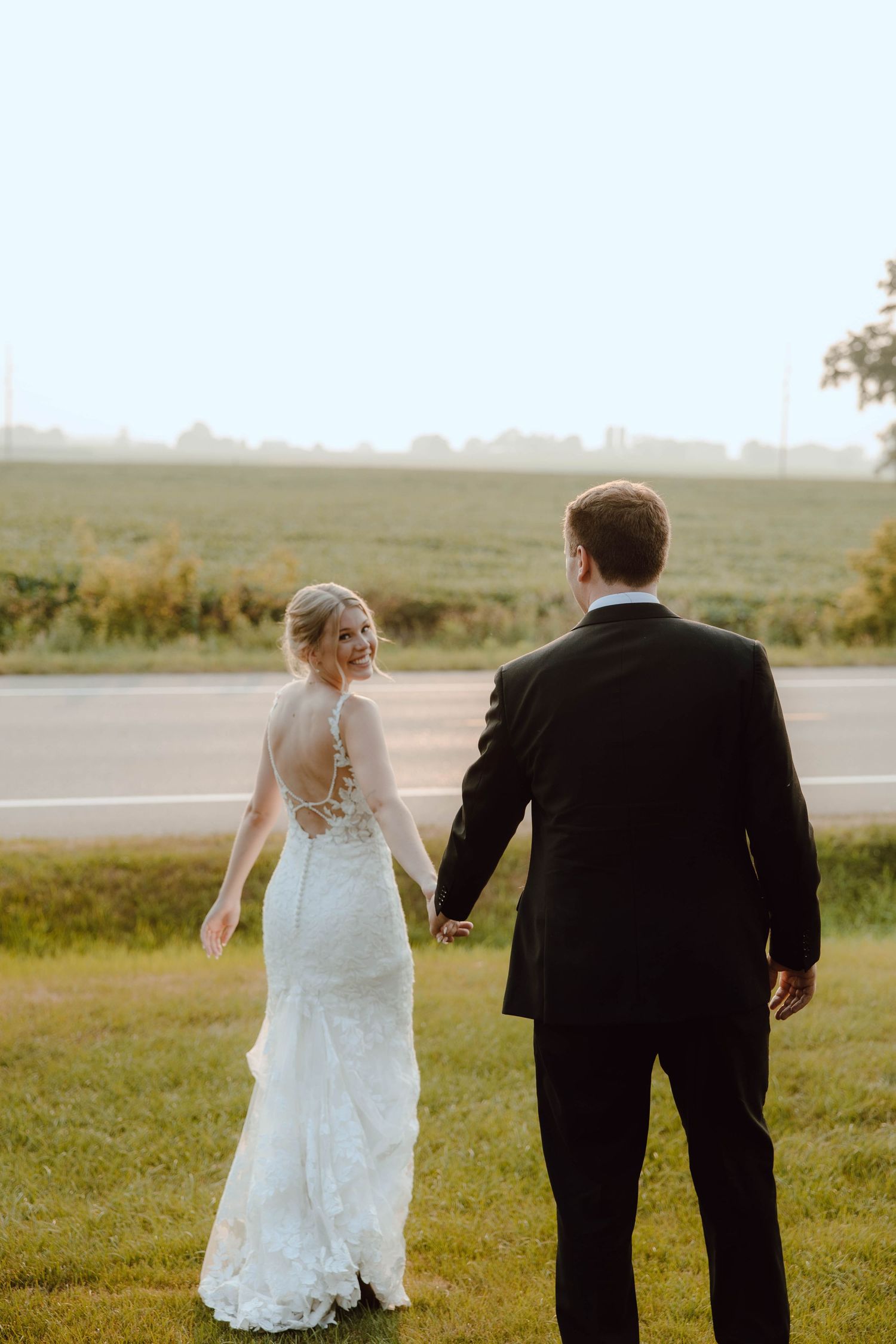 A bride in a lace backless gown holds hands with a groom in a black suit while walking outdoors.