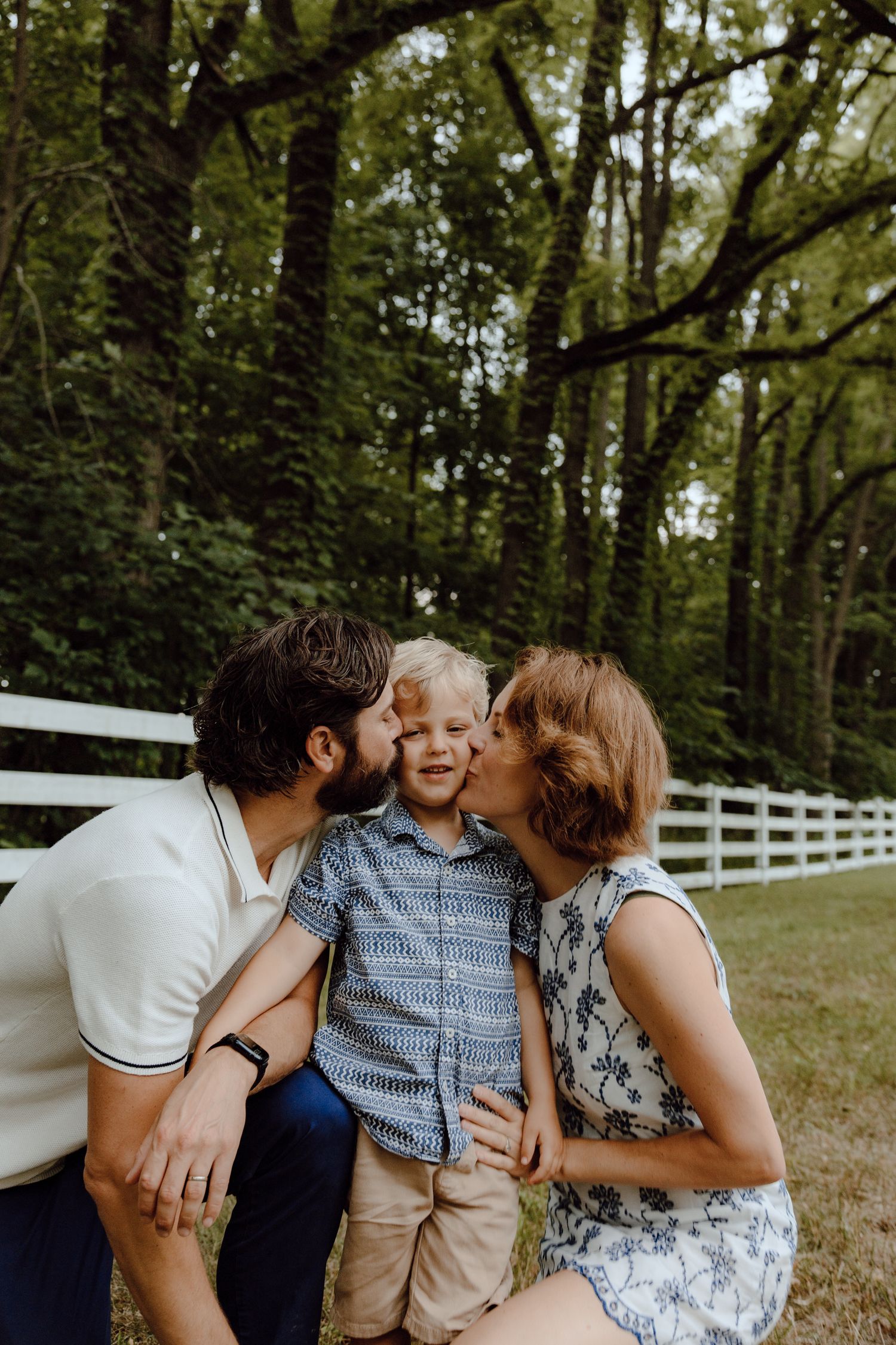 Family sharing affectionate moments together near a white fence in a wooded setting.