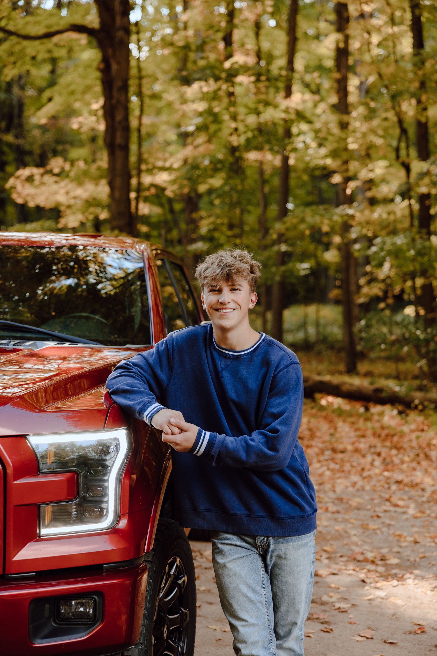A red pickup truck parked in an autumn forest setting with fallen leaves on the ground.