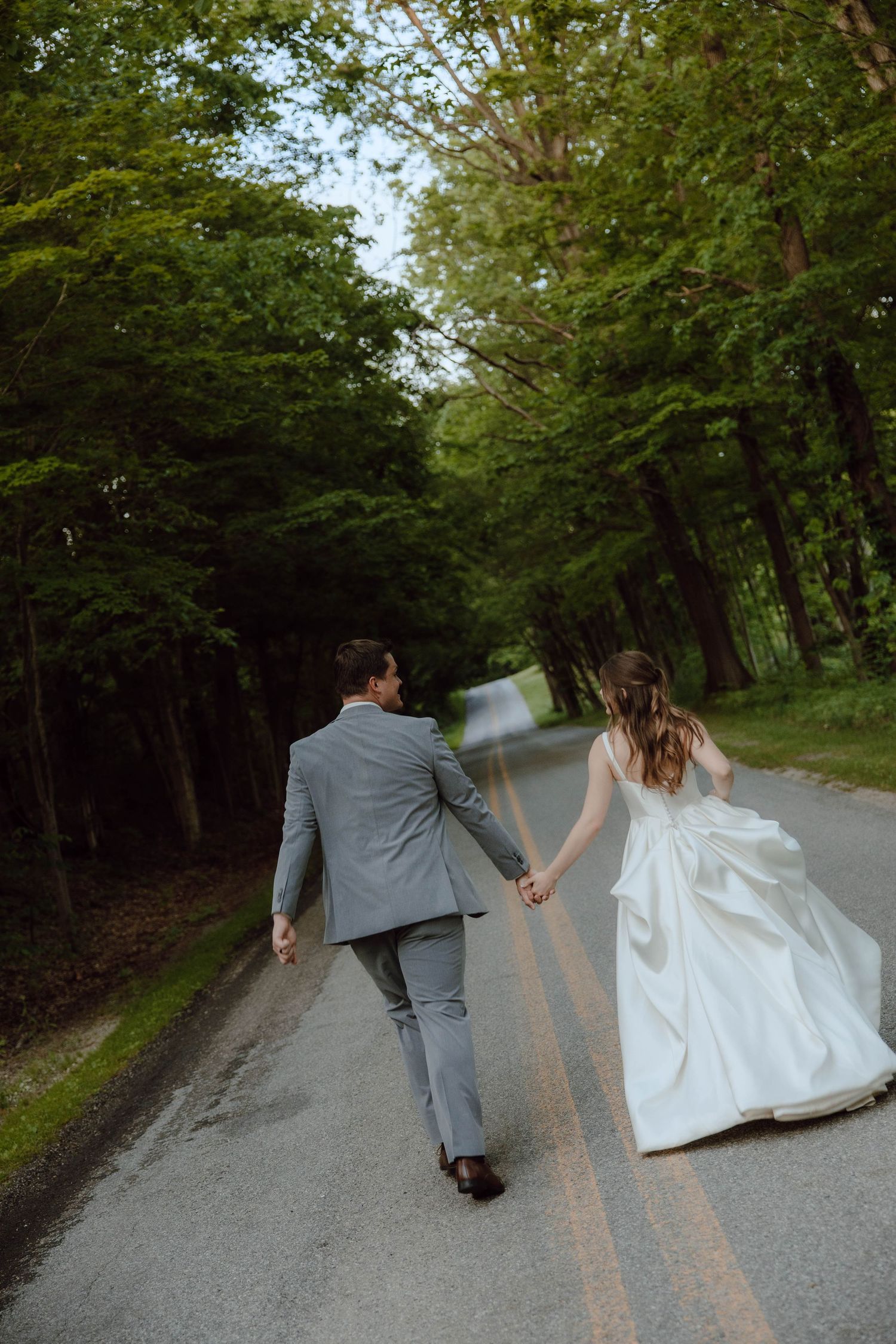 Bride and groom walk hand in hand down a tree-lined road, bride in white gown and groom in gray suit.