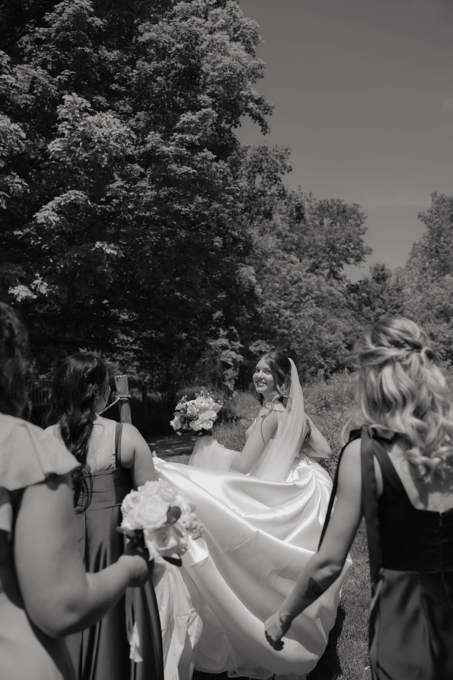 Black and white photo of smiling bride with bridesmaids outdoors, veil flowing in breeze near tall trees.