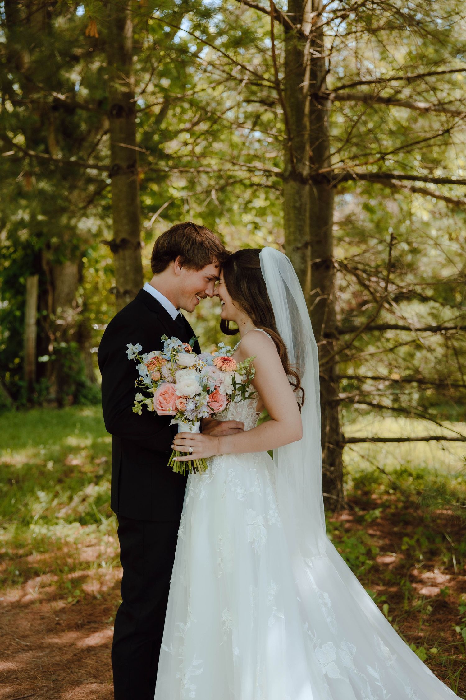 Bride and groom touching foreheads in a forest, bride holding a pink and white bouquet in a lace wedding gown with veil.