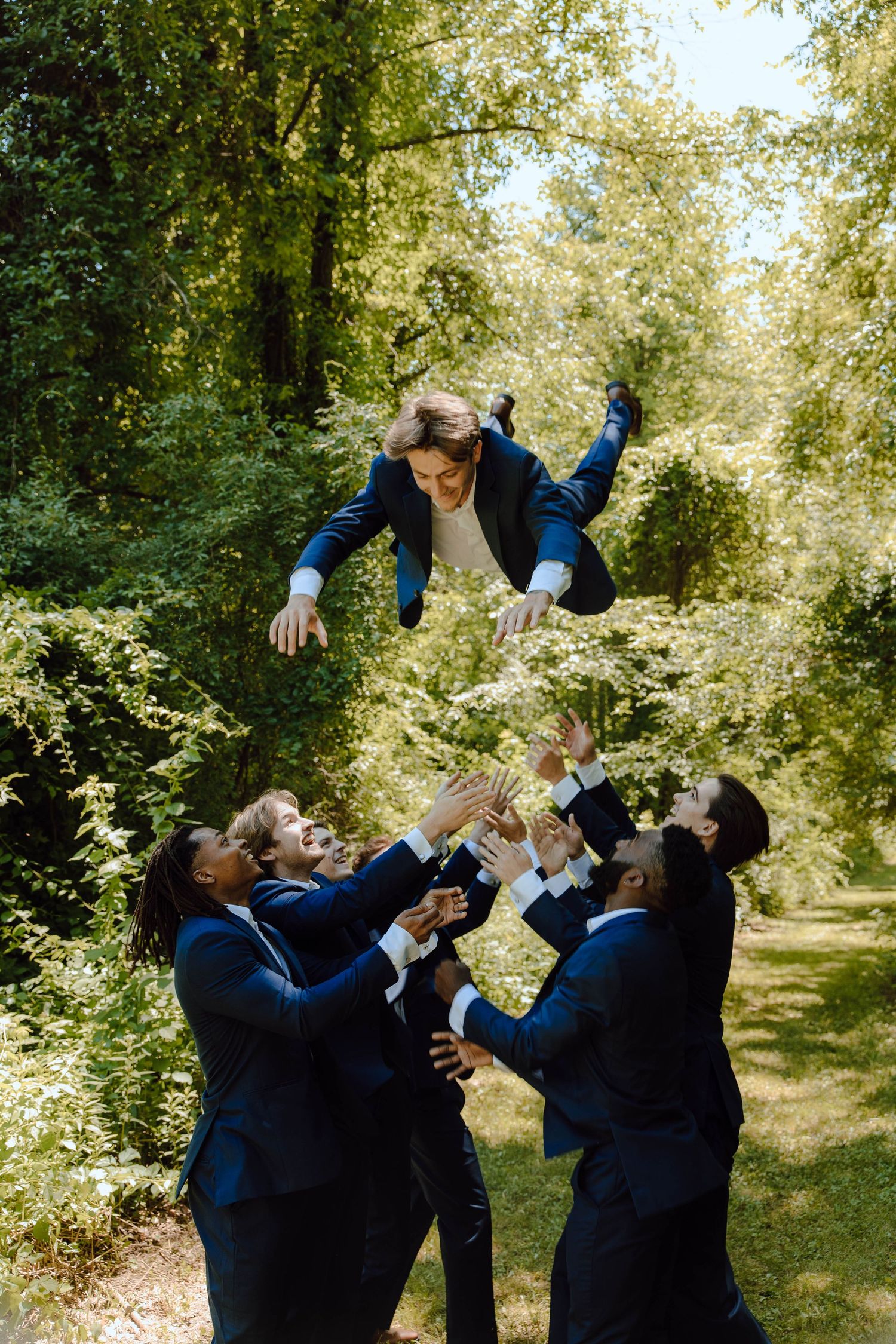 Groomsmen in navy suits tossing the groom into the air outdoors, celebrating at a wedding in a lush green park.