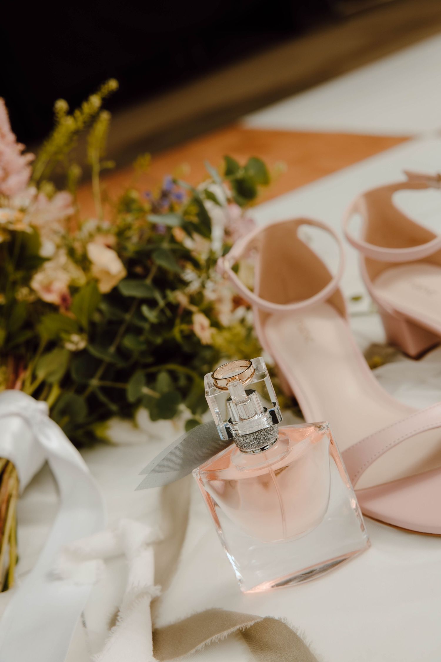 Bridal accessories flat lay with pink perfume bottle, nude heels, and floral bouquet on white surface.