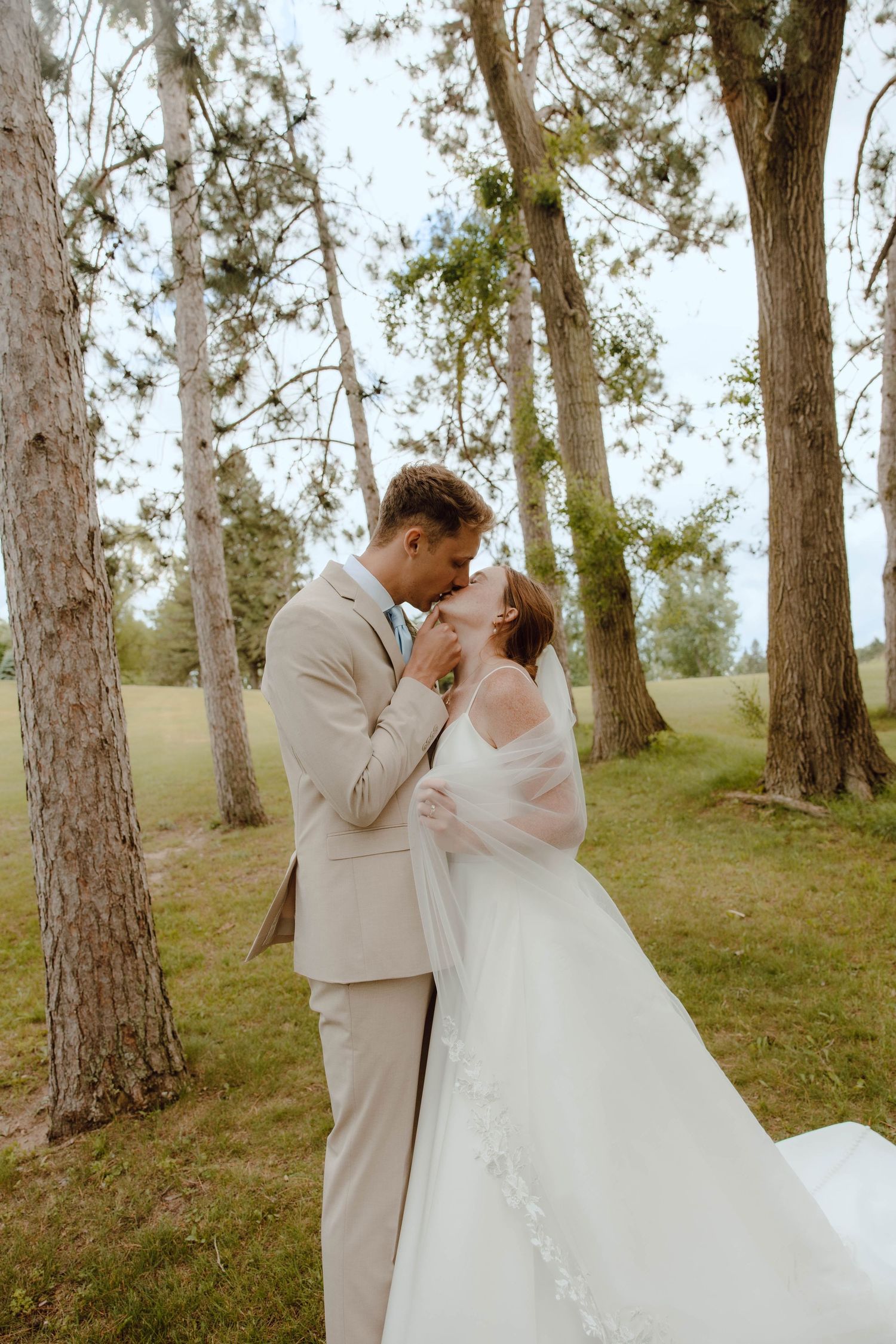 Bride and groom share a romantic kiss among tall trees, she in a flowing white gown with veil, he in a beige suit.