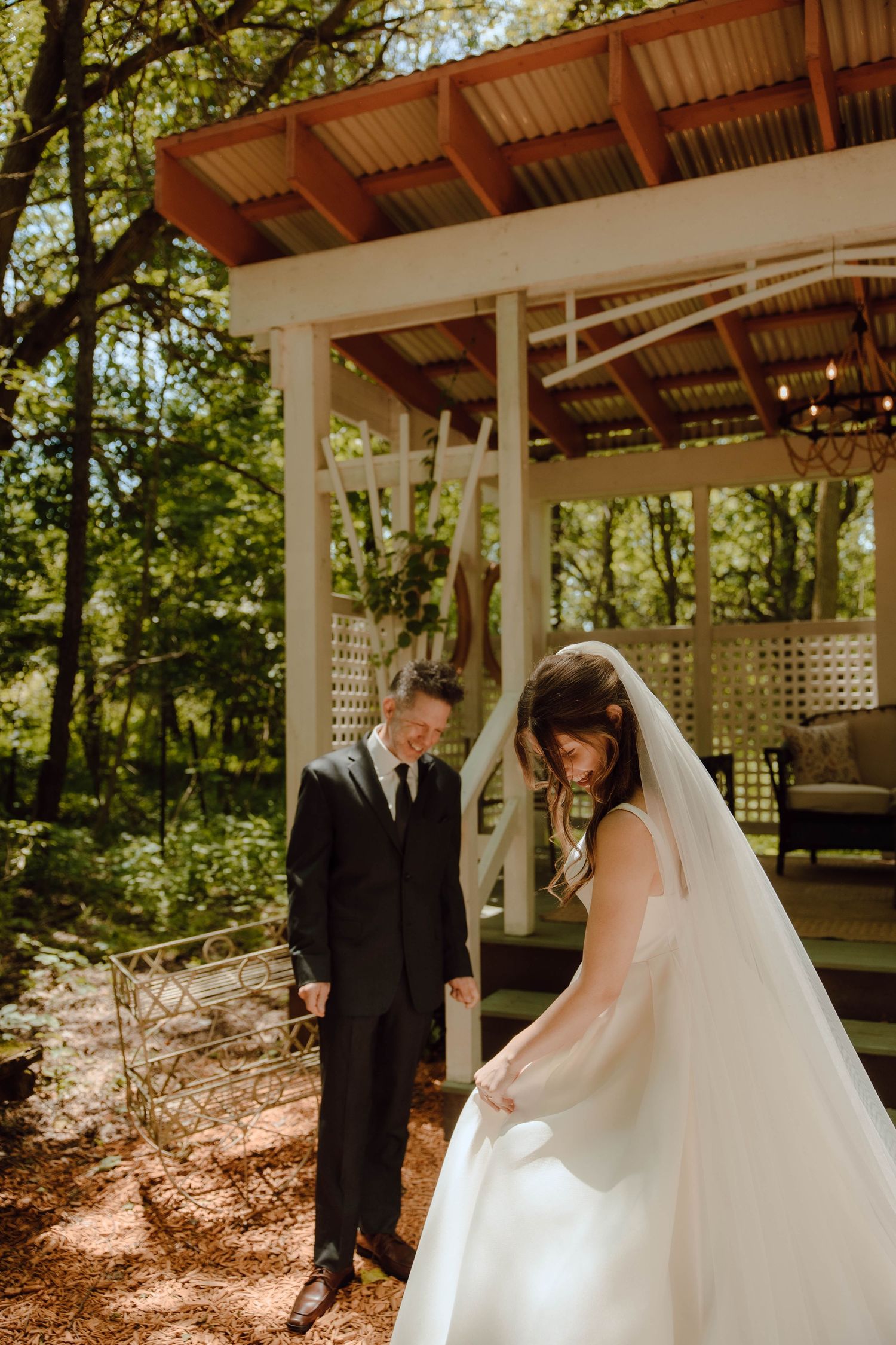 Bride in white gown and groom in black suit share a tender moment outdoors near a rustic gazebo surrounded by trees.
