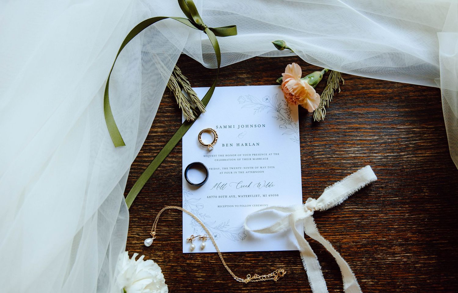Wedding flat lay with invitation, gold rings, bridal veil, floral accents, and jewelry on rustic wood.