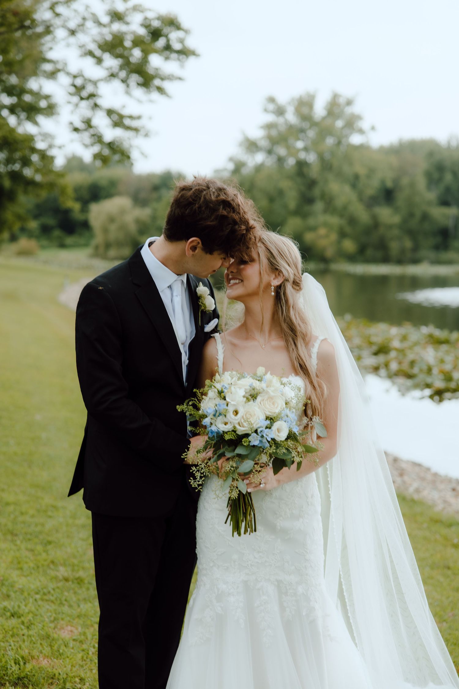 Bride and groom share a romantic forehead touch outdoors near a pond, bride holding a white and blue bouquet.