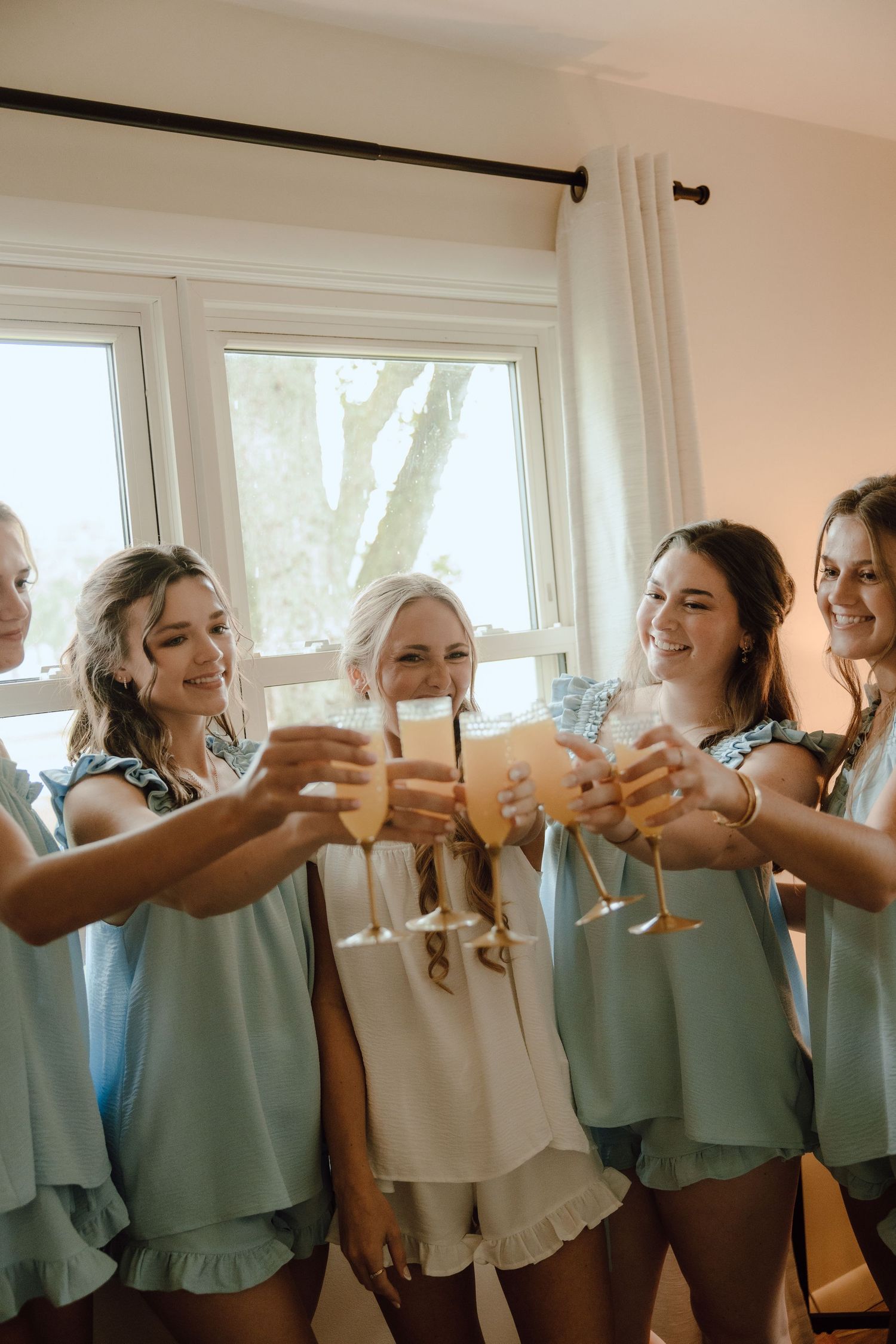 Bride and bridesmaids in sage robes toasting with champagne glasses during wedding morning preparations.