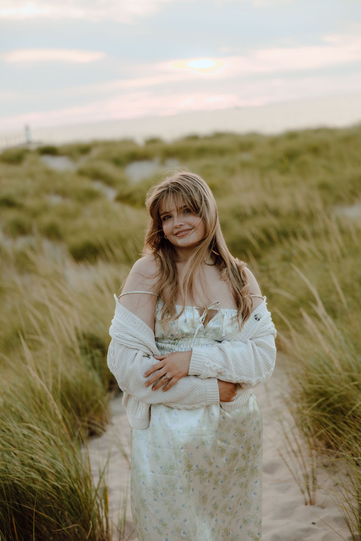 Young blonde woman in floral dress and white jacket smiling on sandy beach dunes at sunset.