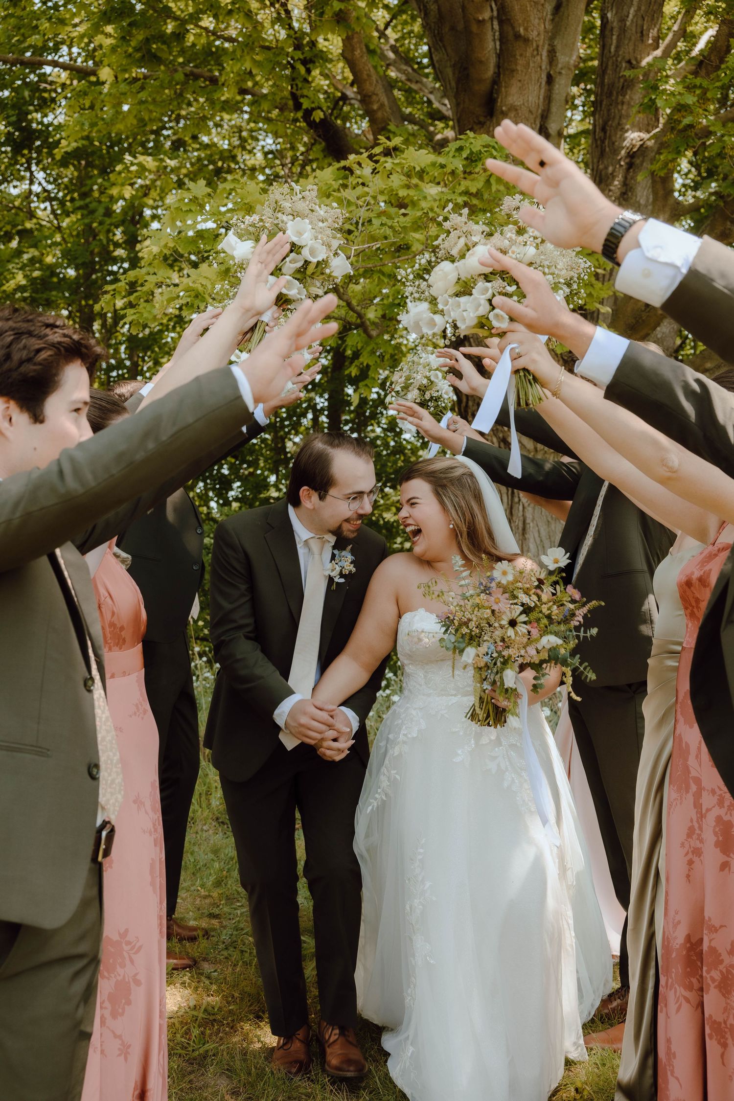 Newlyweds walk hand-in-hand under an arch of raised hands formed by their wedding party outdoors.