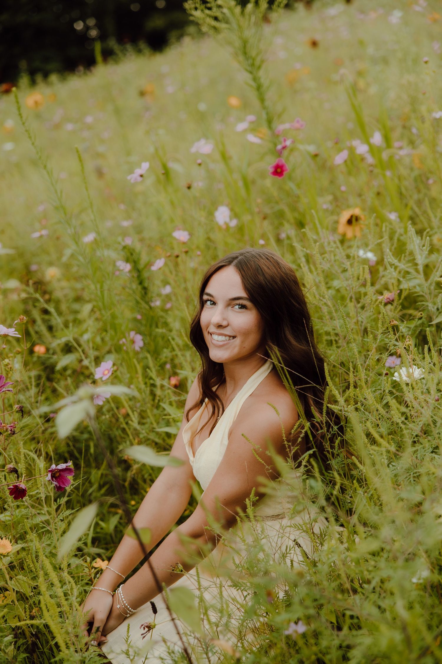 Young woman in yellow dress sitting among colorful wildflowers in a lush green meadow, smiling warmly.