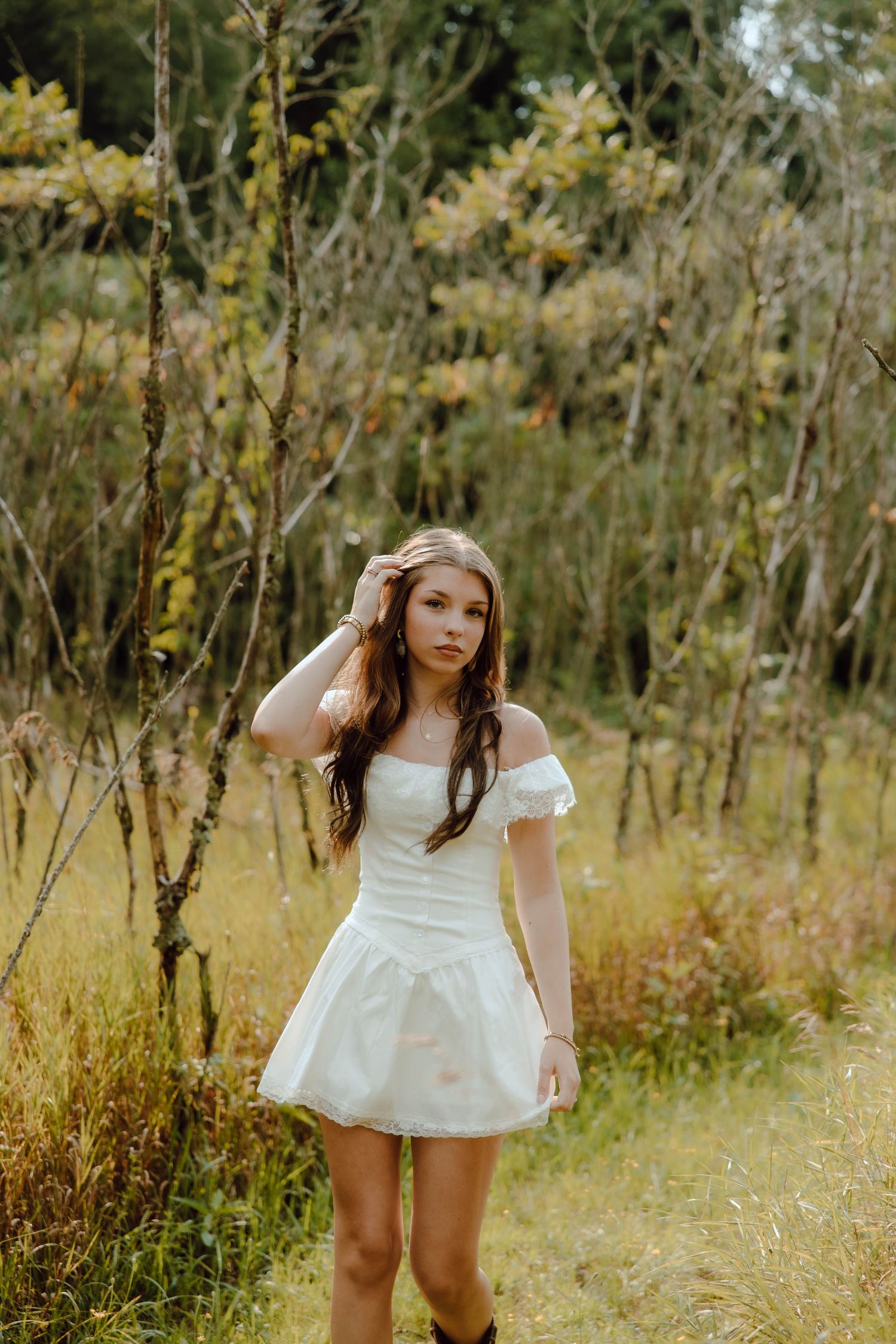 Young woman in white off-shoulder mini dress posing in a forest meadow with tall grass and trees.