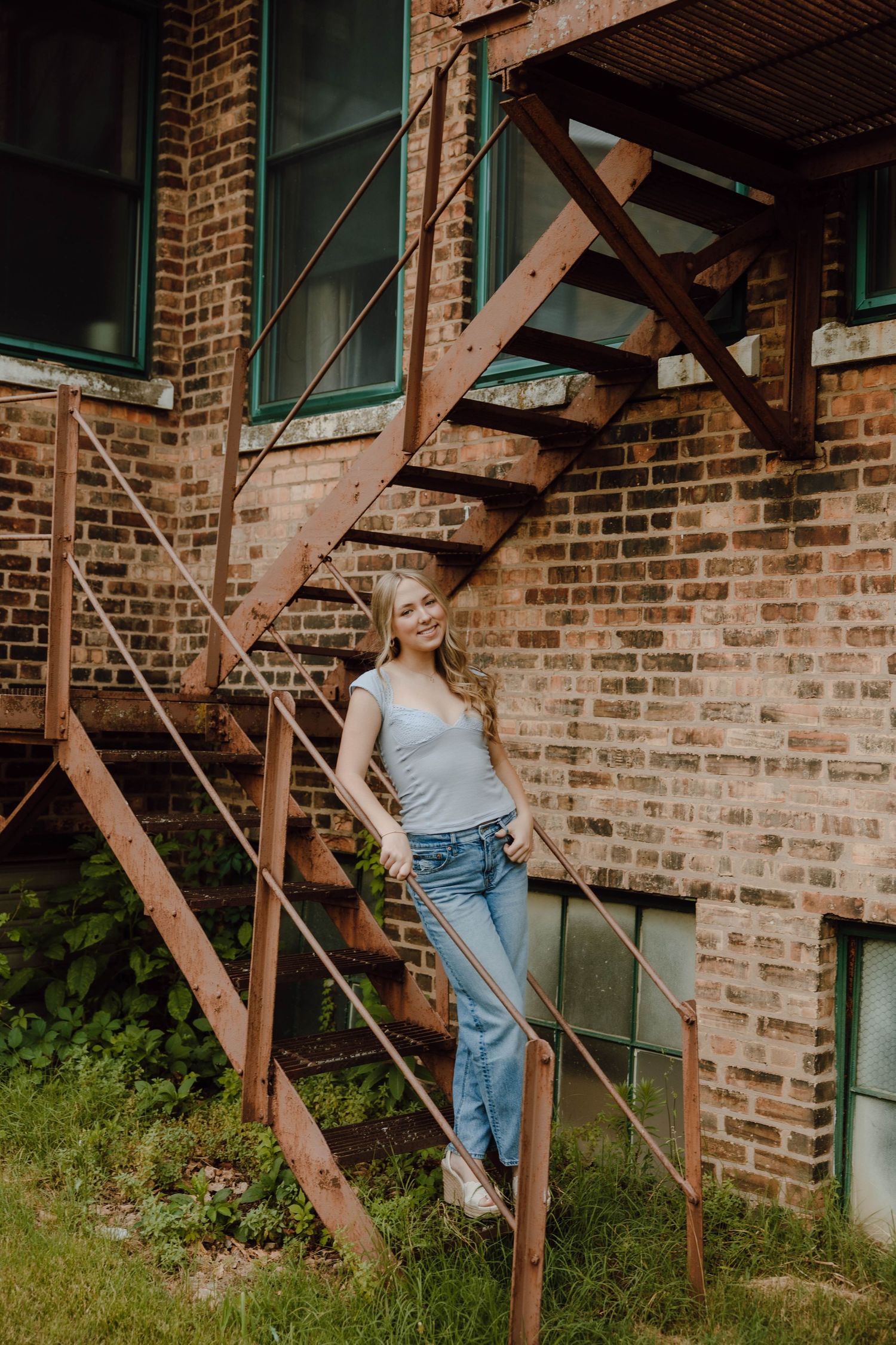 Young woman in jeans and gray top posing on rustic metal fire escape stairs against brick building.