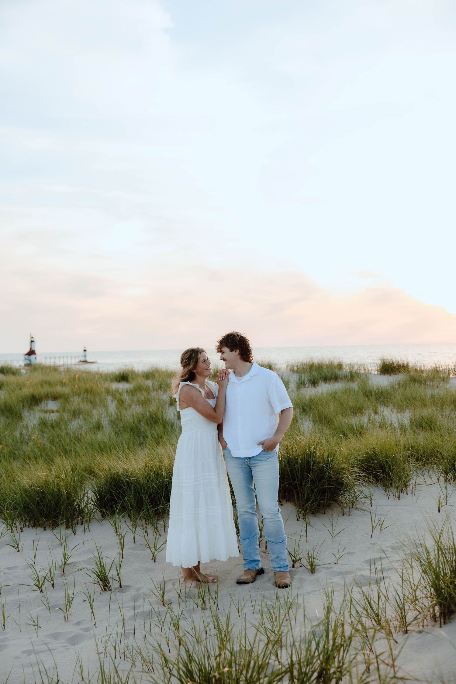 Couple sharing a romantic moment on sandy beach dunes at sunset, woman in white dress, man in jeans.