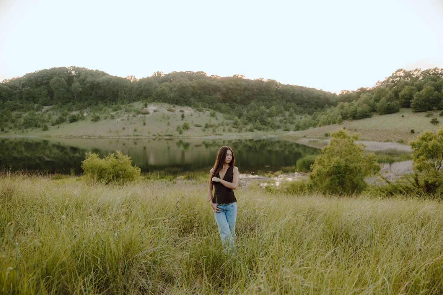 Young woman standing in tall grass meadow with scenic lake and green hills in background.