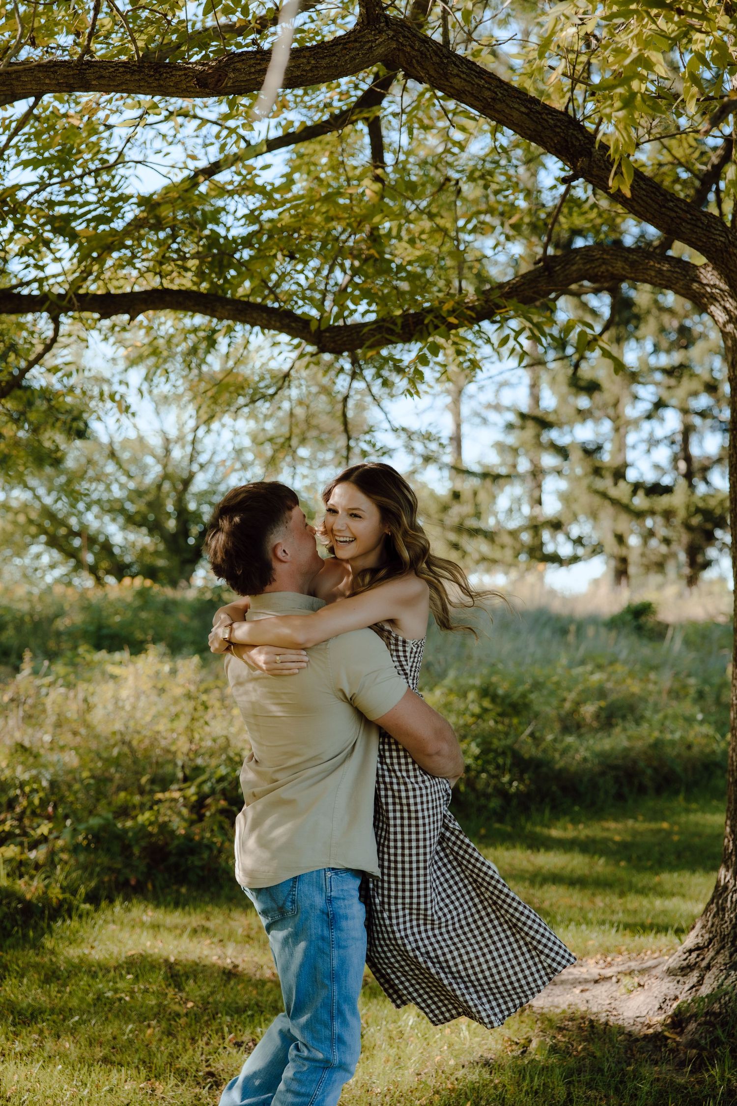 A joyful couple embraces and spins under a tree in a lush green park on a sunny day.