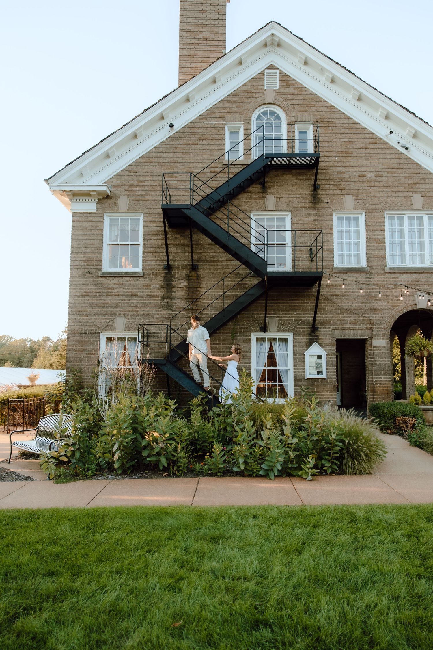 Couple in wedding attire descends black iron staircase on brick building exterior surrounded by lush greenery.