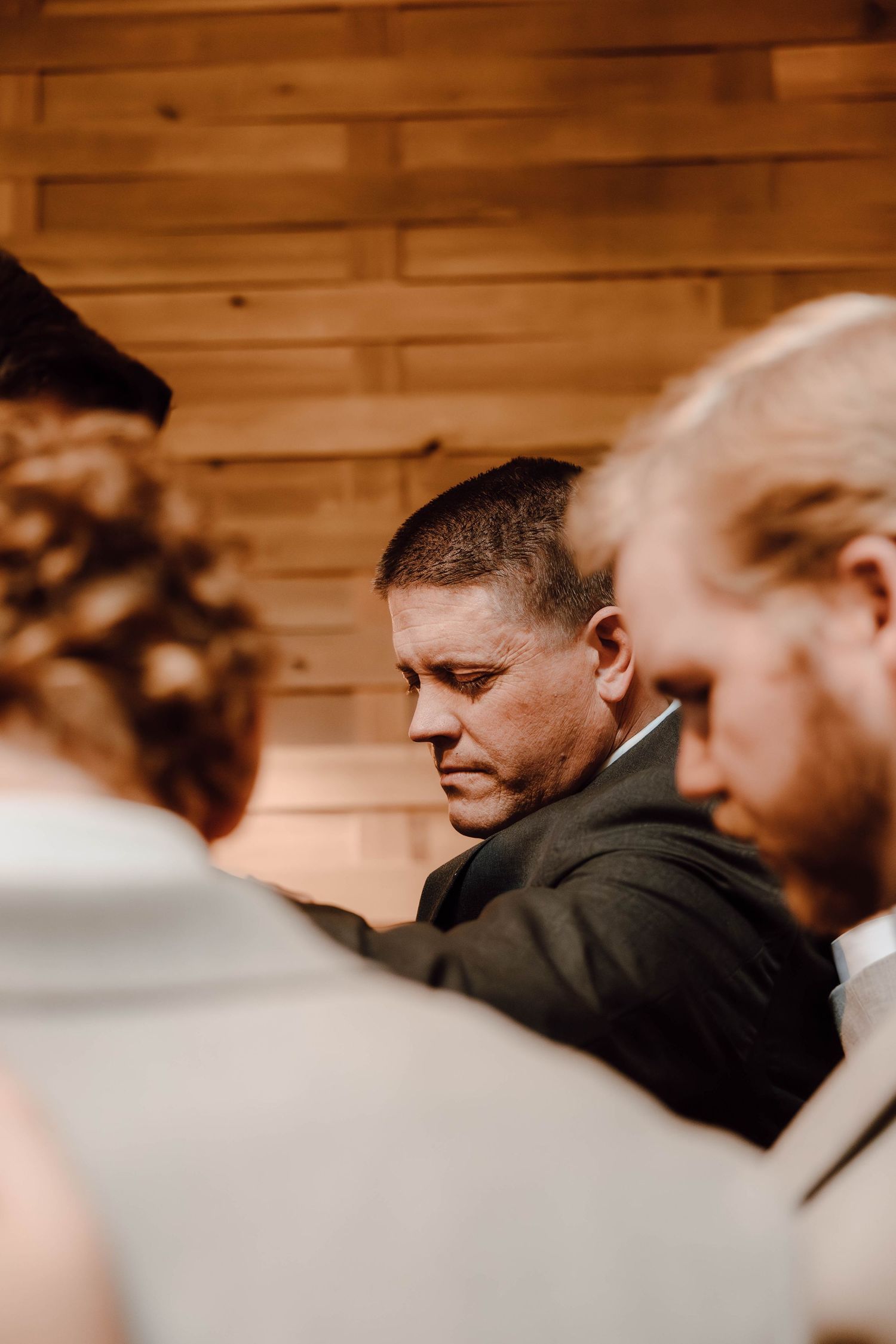 Wedding guests bow heads in prayer inside rustic wood-paneled venue during ceremony.
