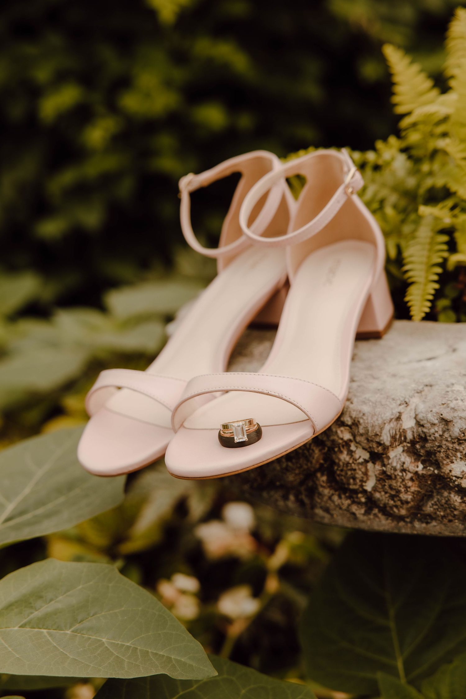 Blush pink block heel sandals with gold wedding rings displayed on mossy stone surrounded by ferns.